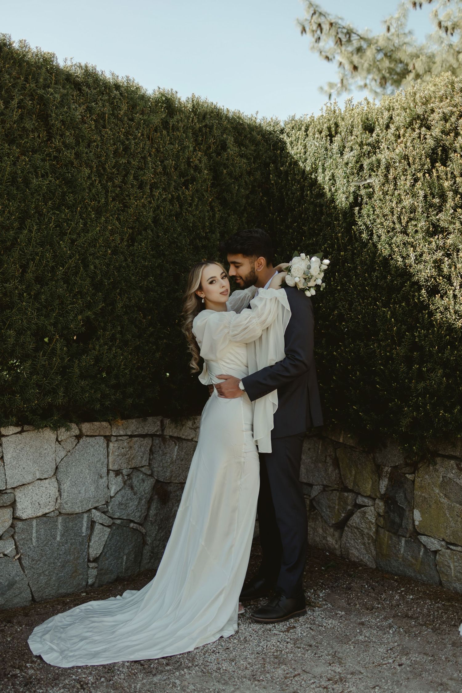 Romantic wedding couple embracing against a hedge wall in elegant formal attire.