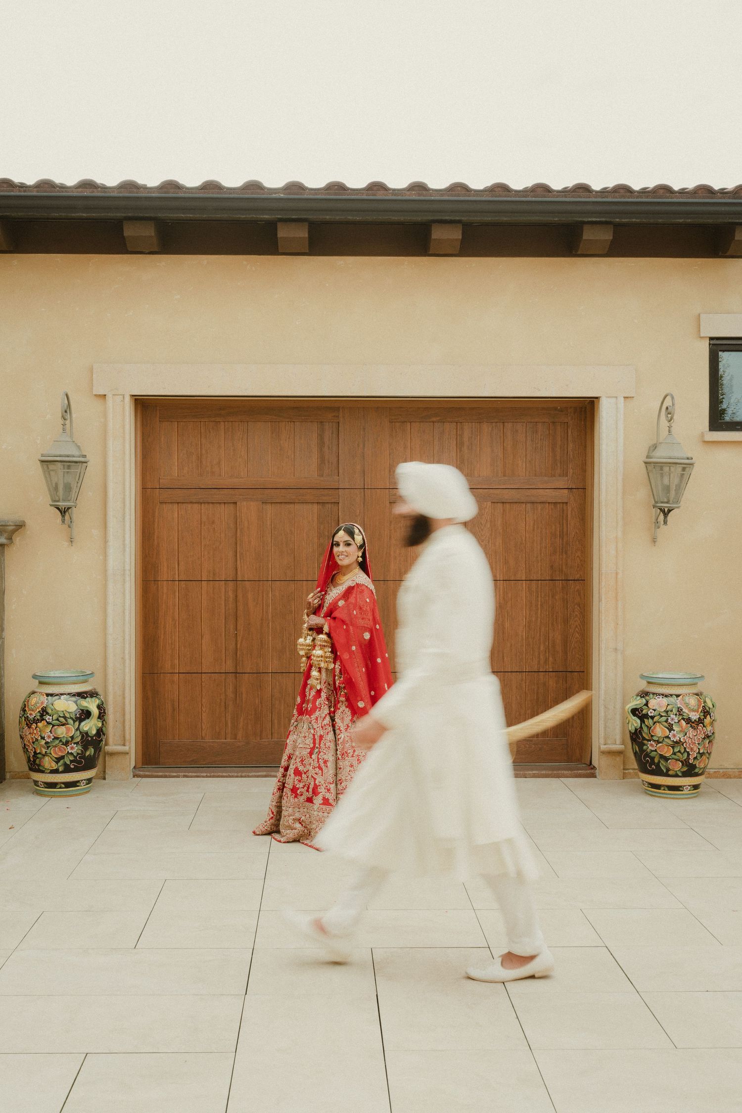 Bride in red lehenga stands by wooden doors as groom in white sherwani walks past during Indian wedding.