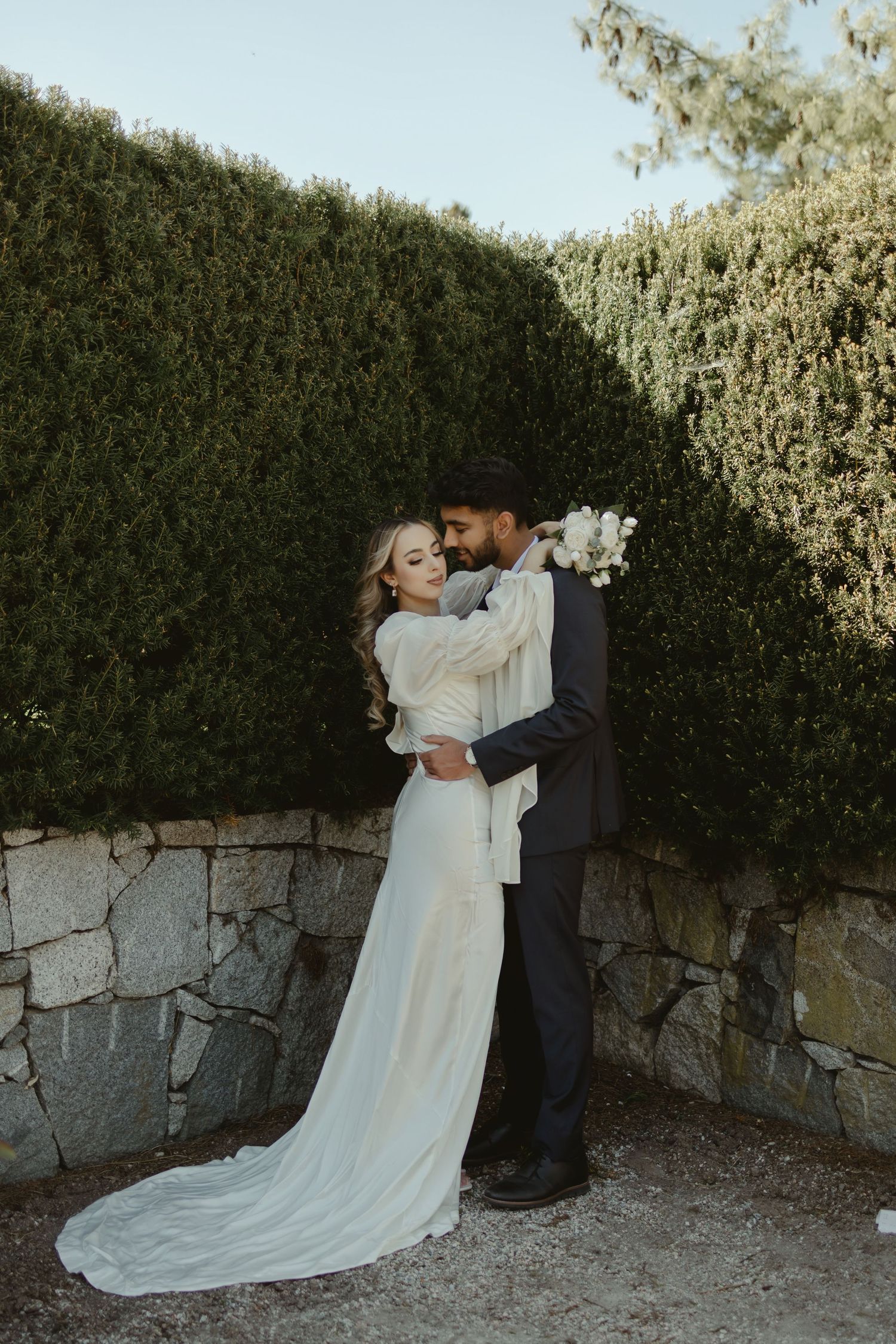 Romantic couple embrace against hedge wall with flowing white wedding dress.