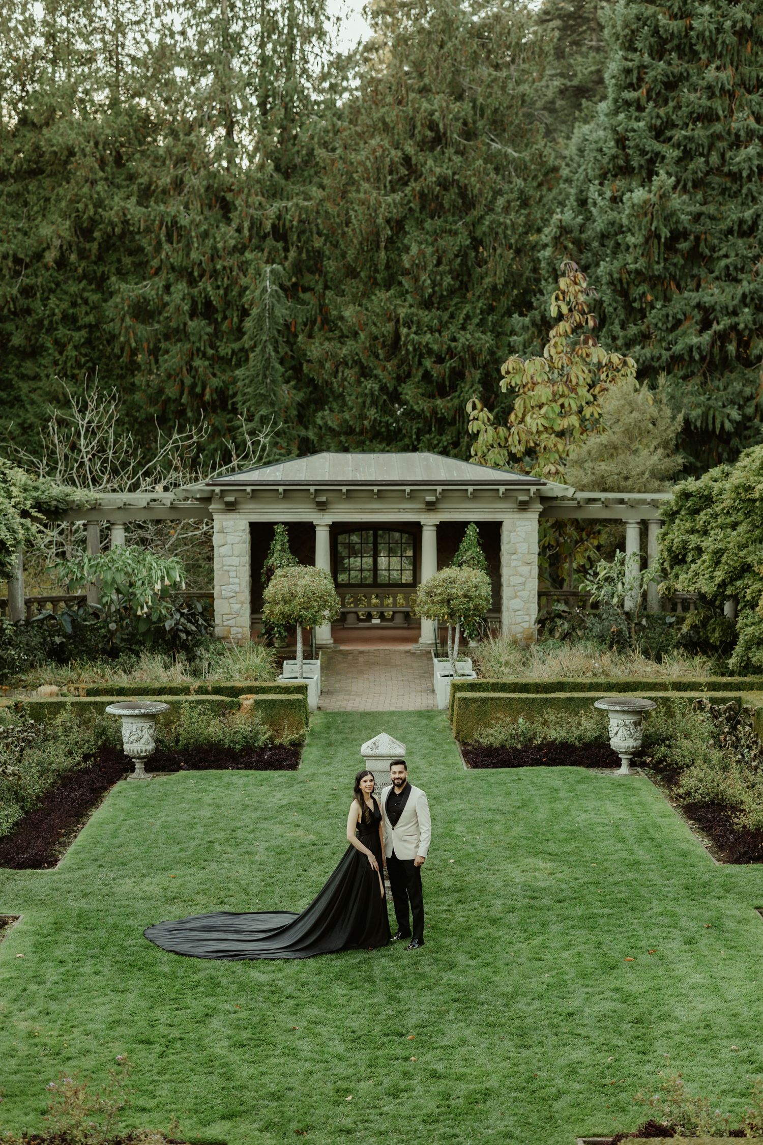 A romantic outdoor scene in a formal garden with manicured lawn and stone pavilion featuring two figures in elegant attire.