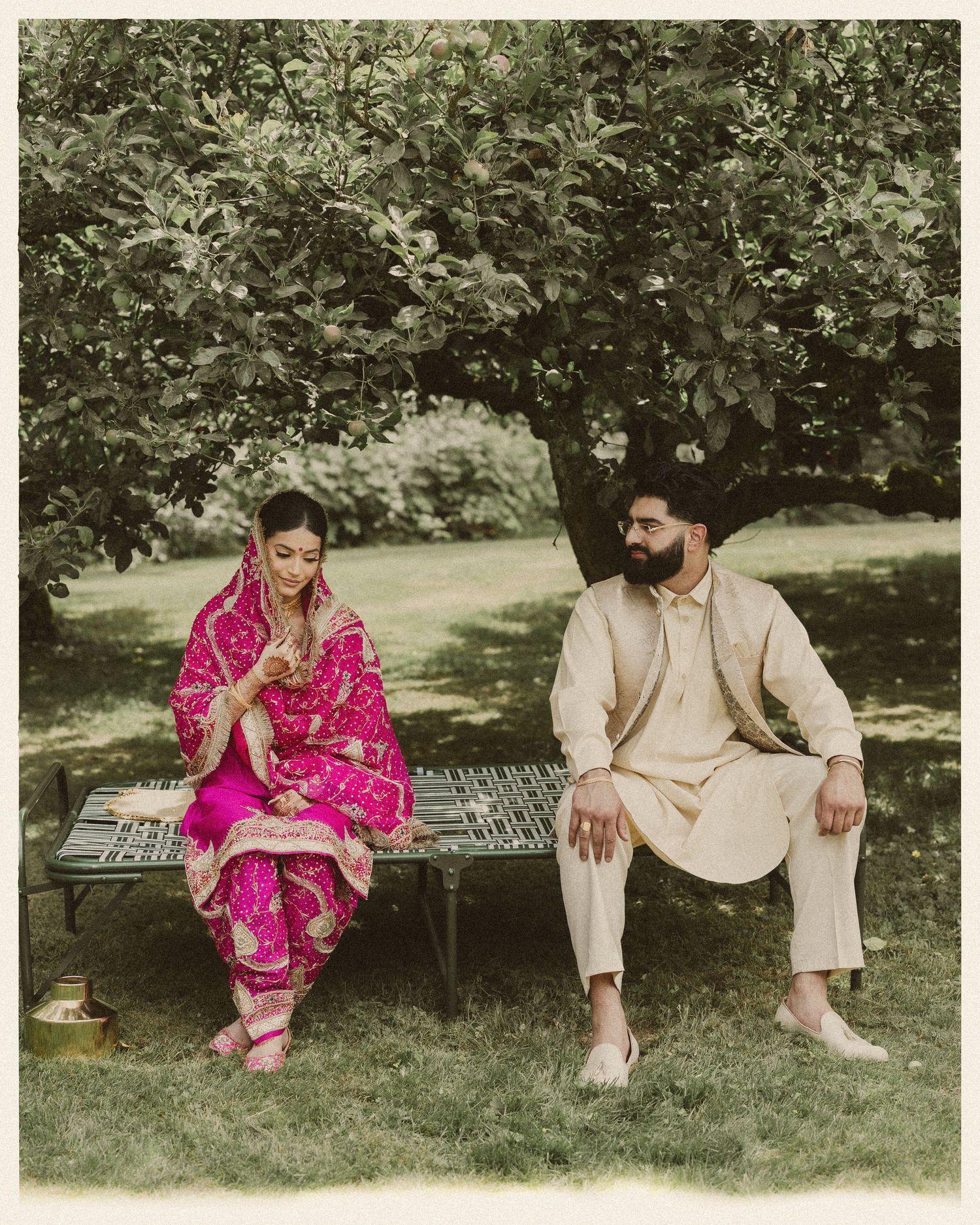 Couple in traditional South Asian attire sitting on garden bench under tree.
