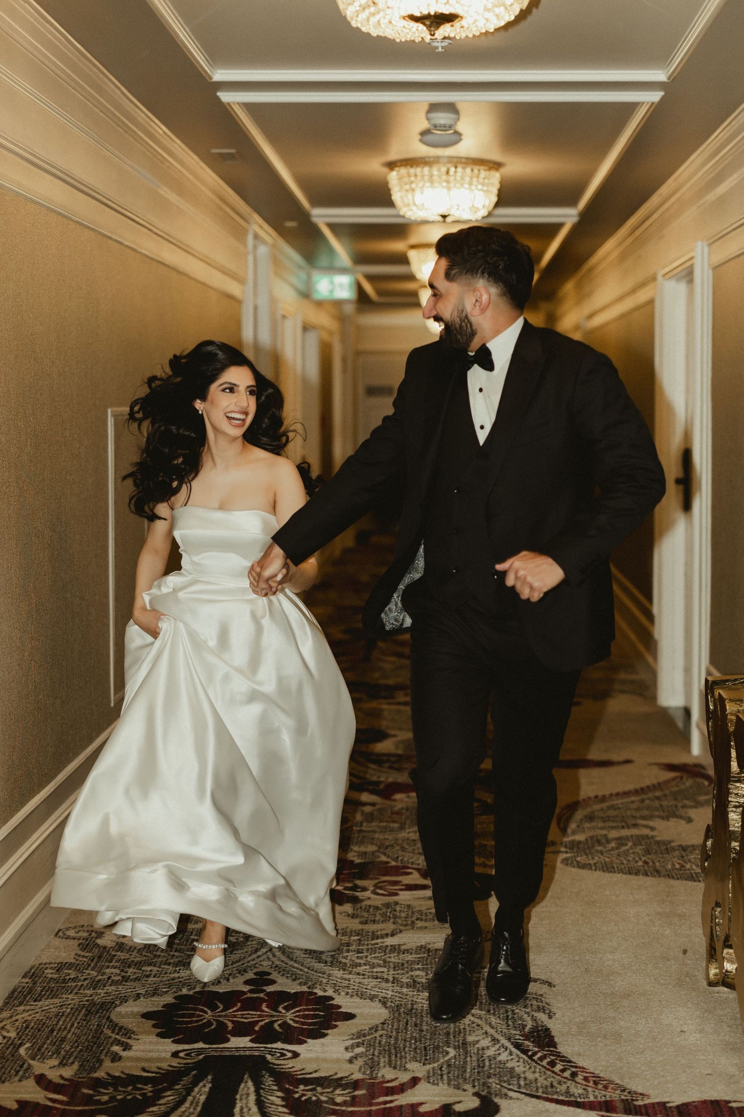 A couple runs playfully down an elegant hotel hallway in formal wedding attire.