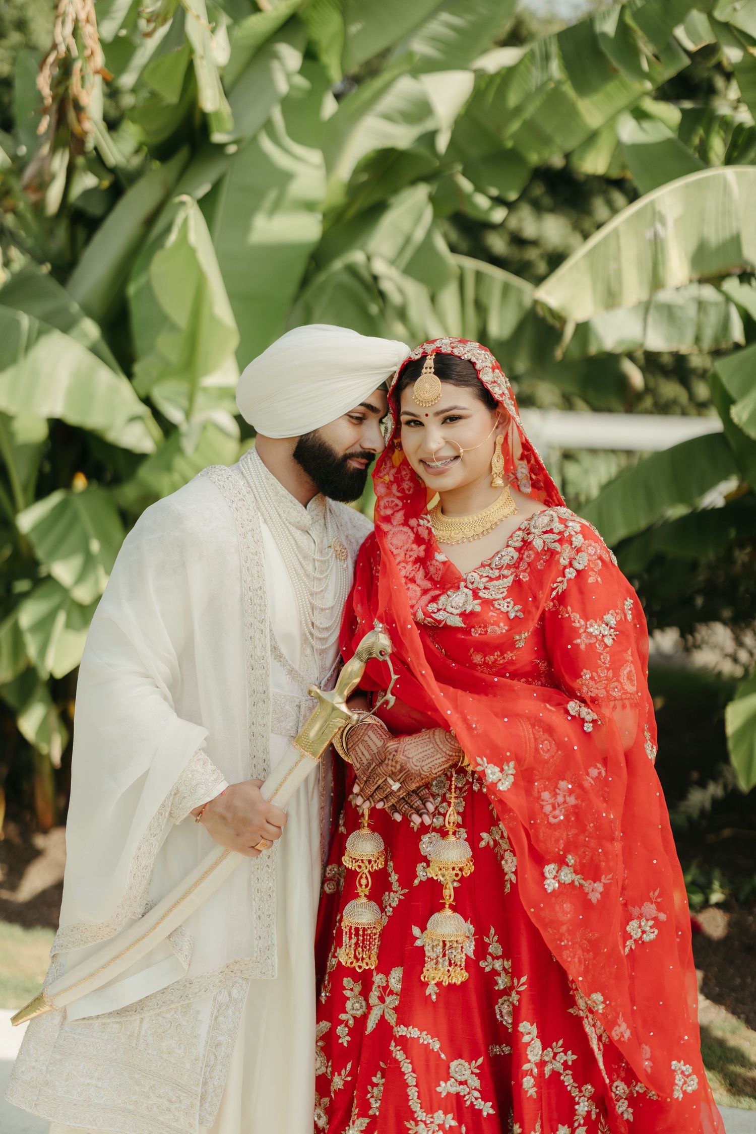 Traditional Indian wedding couple in vibrant red and white attire pose together against lush green foliage background.