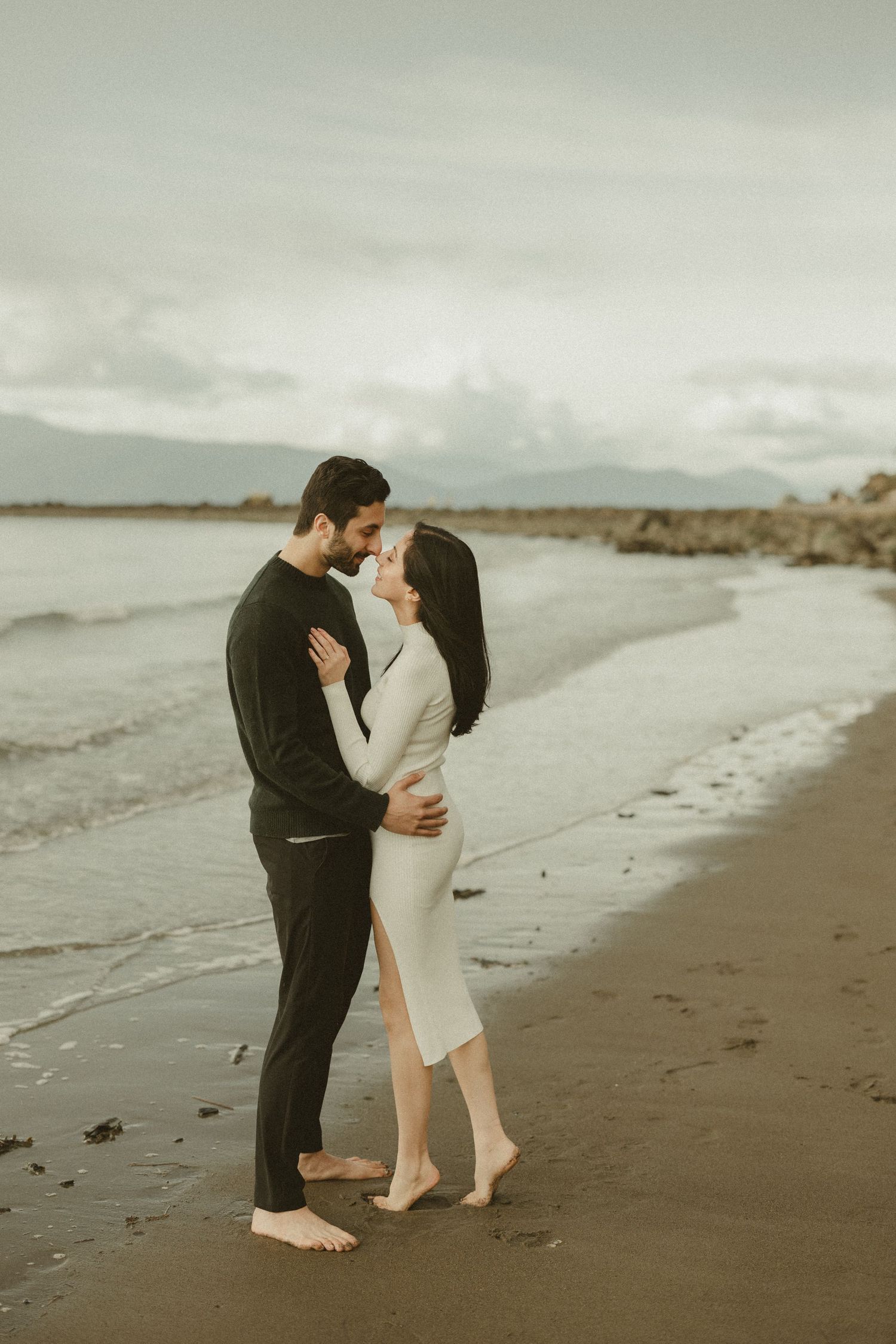 Couple sharing an intimate moment on a cloudy beach in casual attire.