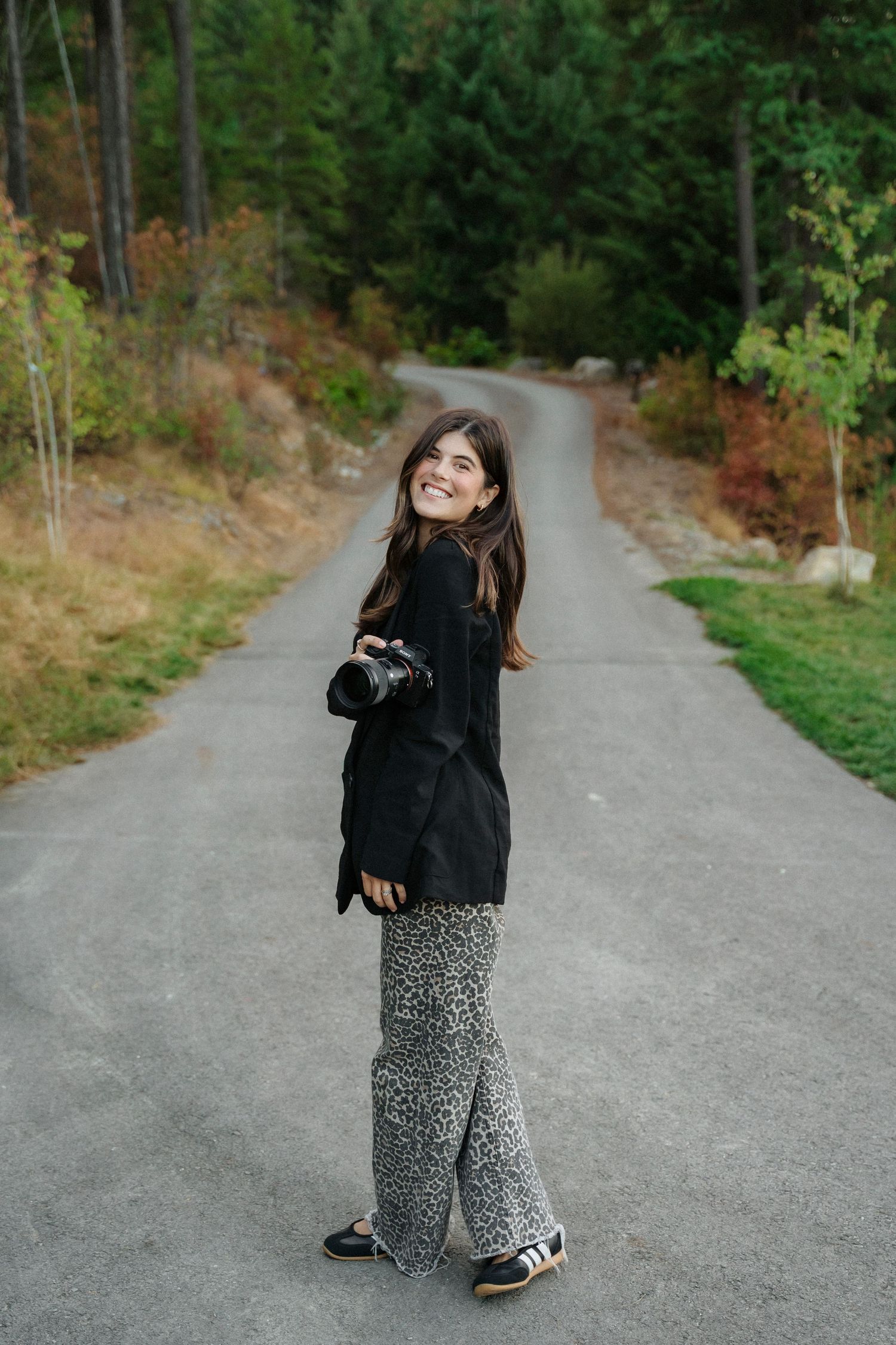 Smiling woman holding a camera stands on a forest road wearing a black jacket and leopard print wide-leg pants.