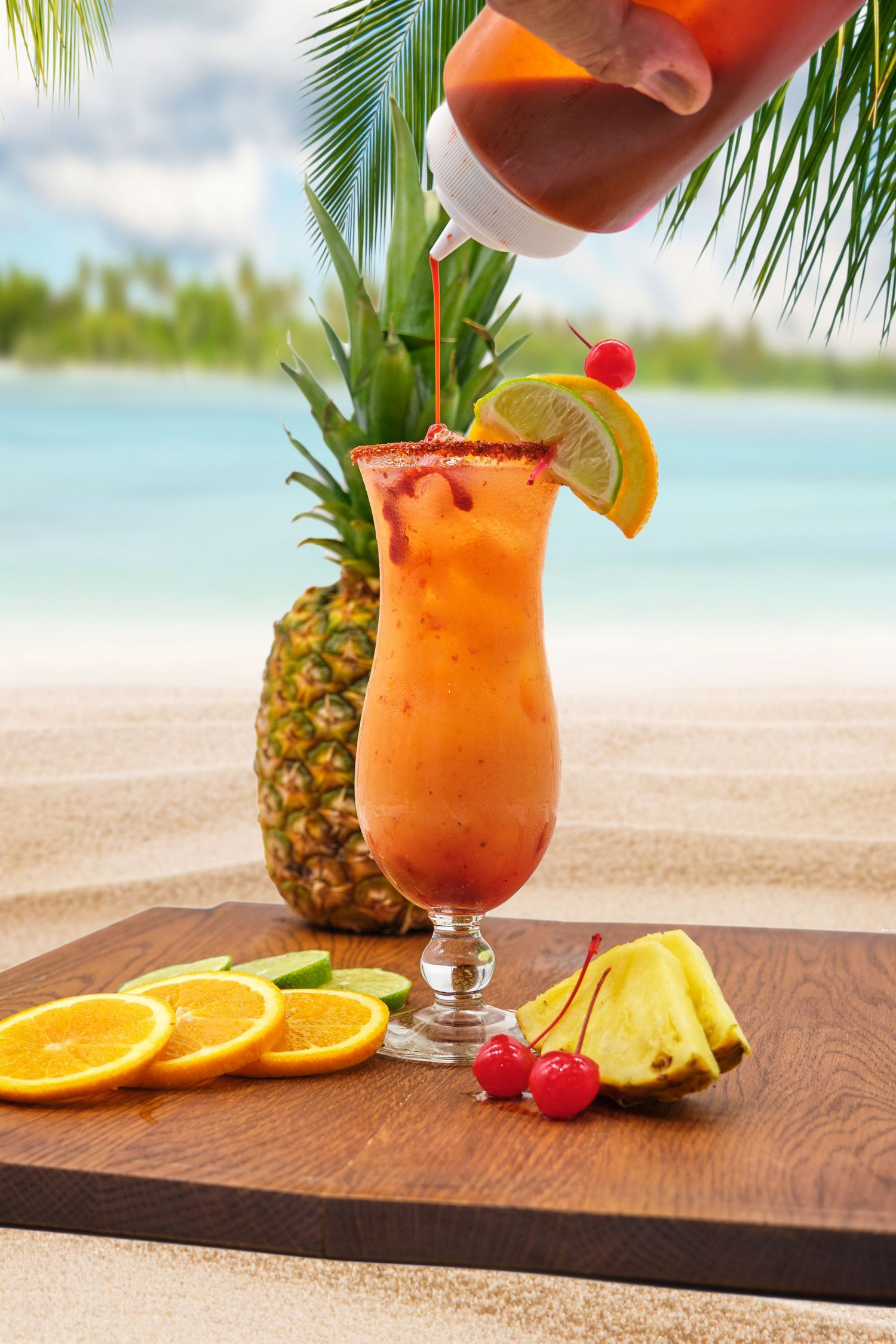 Tropical cocktail being poured on beach with pineapple and orange garnish.