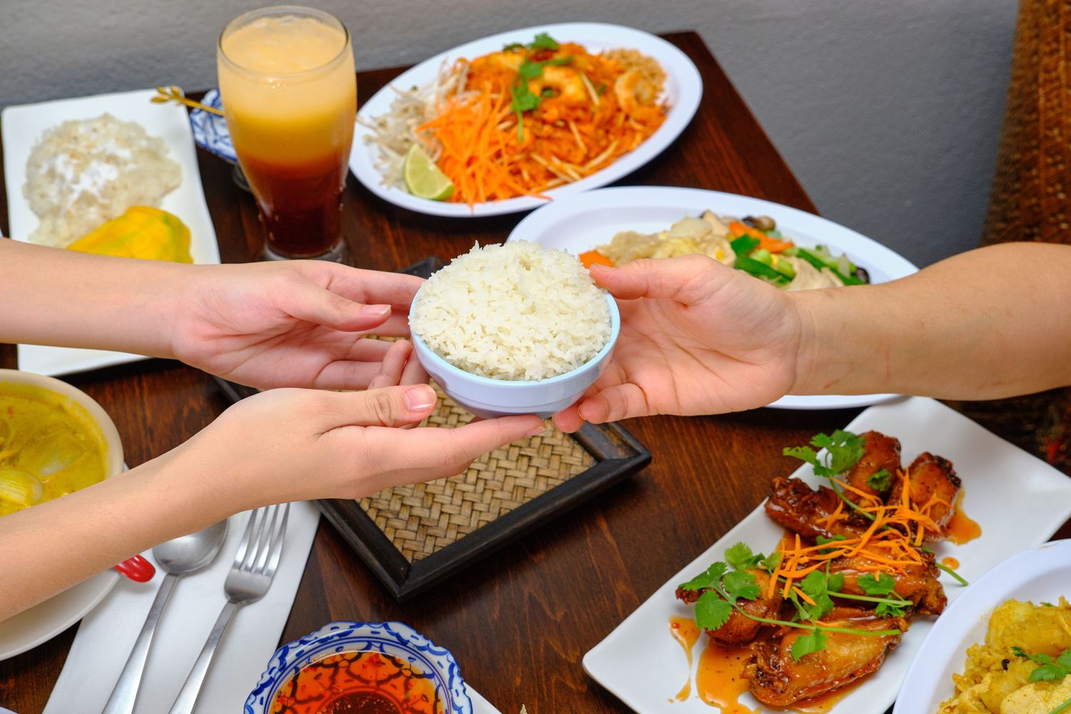 Hands passing rice bowls across a table with various Asian dishes and beverages.