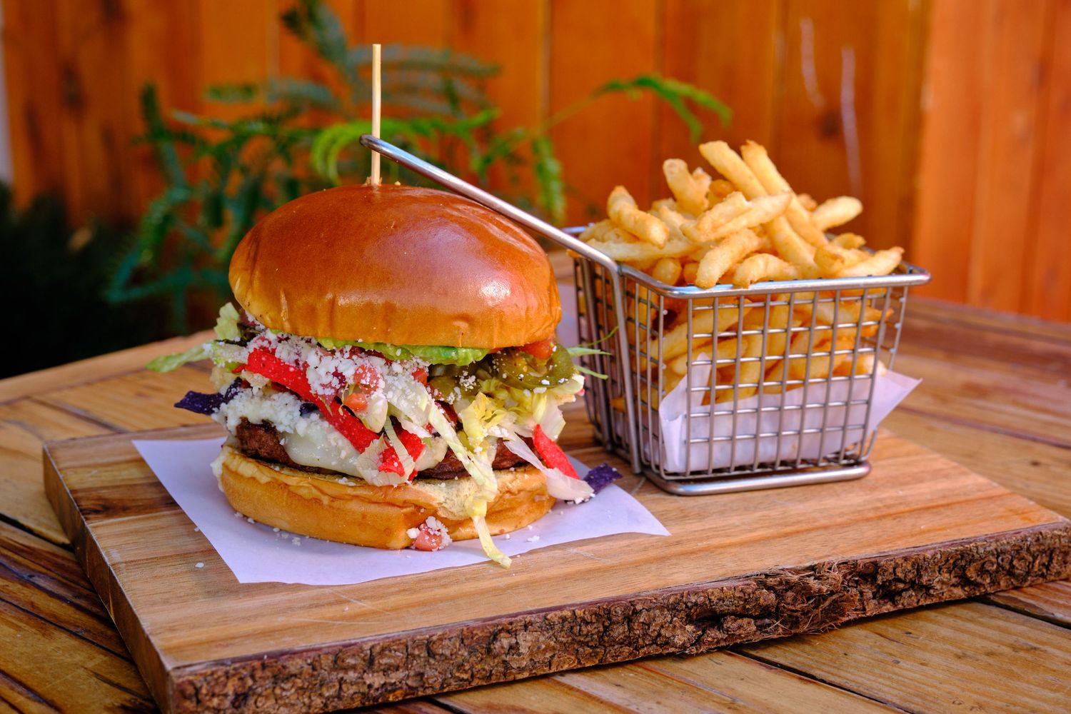 Juicy burger with toppings served on wooden board with crispy french fries in metal basket.