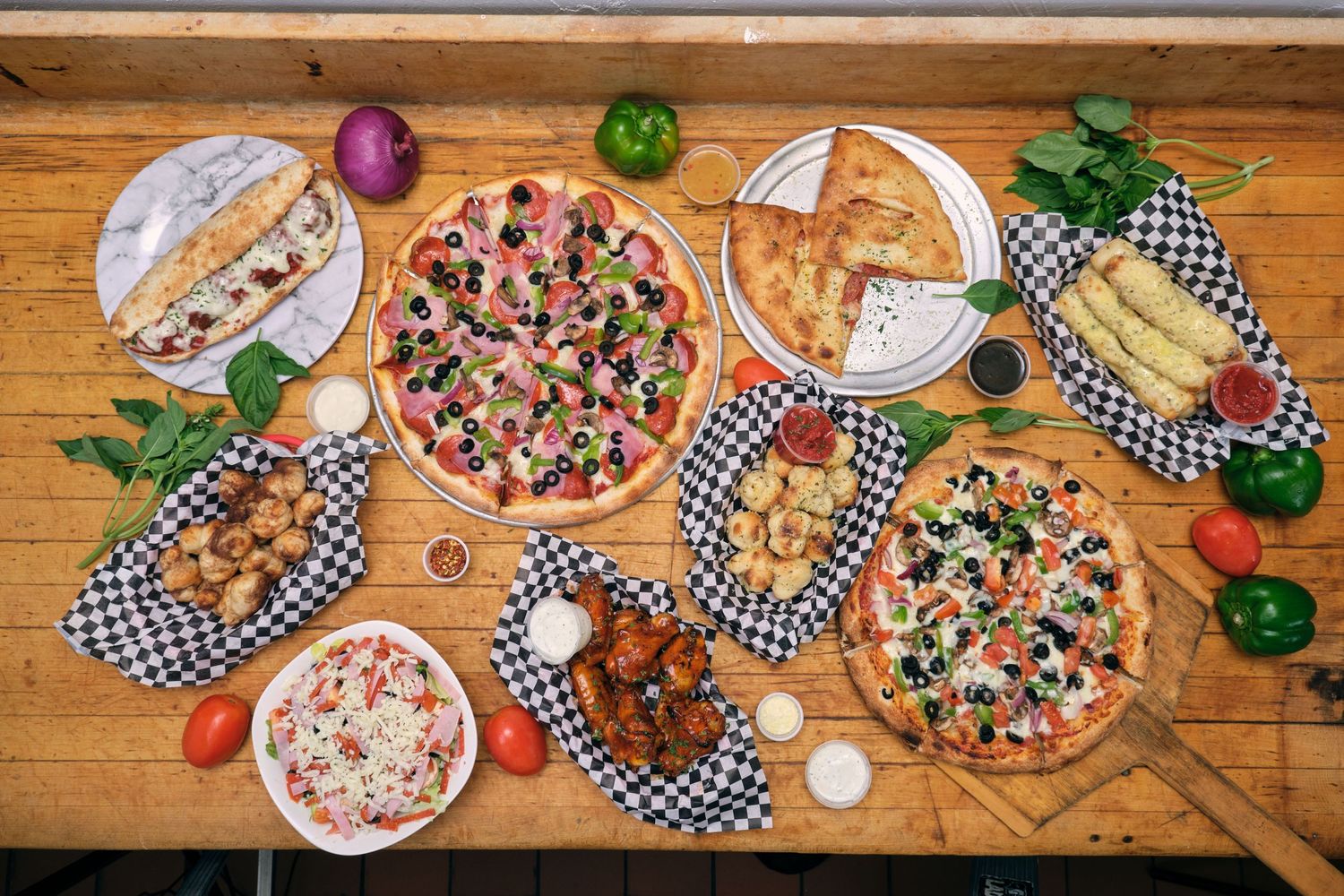 Birds-eye view of various pizzas and appetizers arranged on wooden table with checkered liners.
