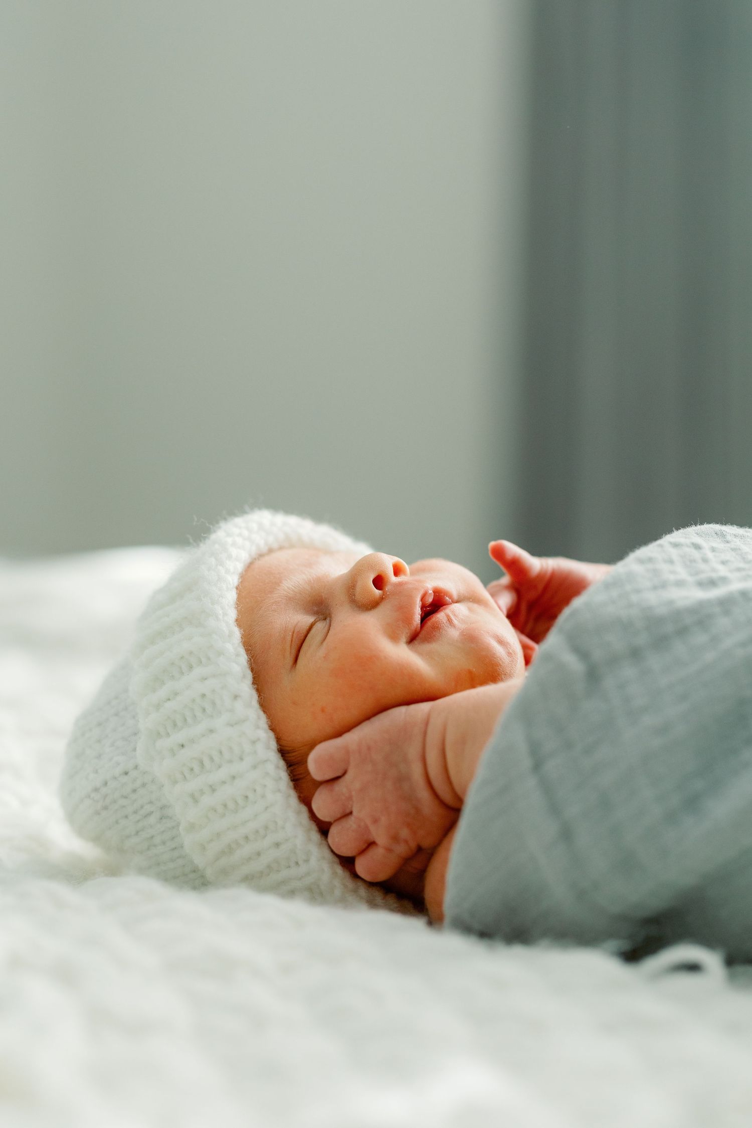 A newborn lies, swaddled in a light green blanket holding their hand up to their cheeks. Photo by Katie Crane.