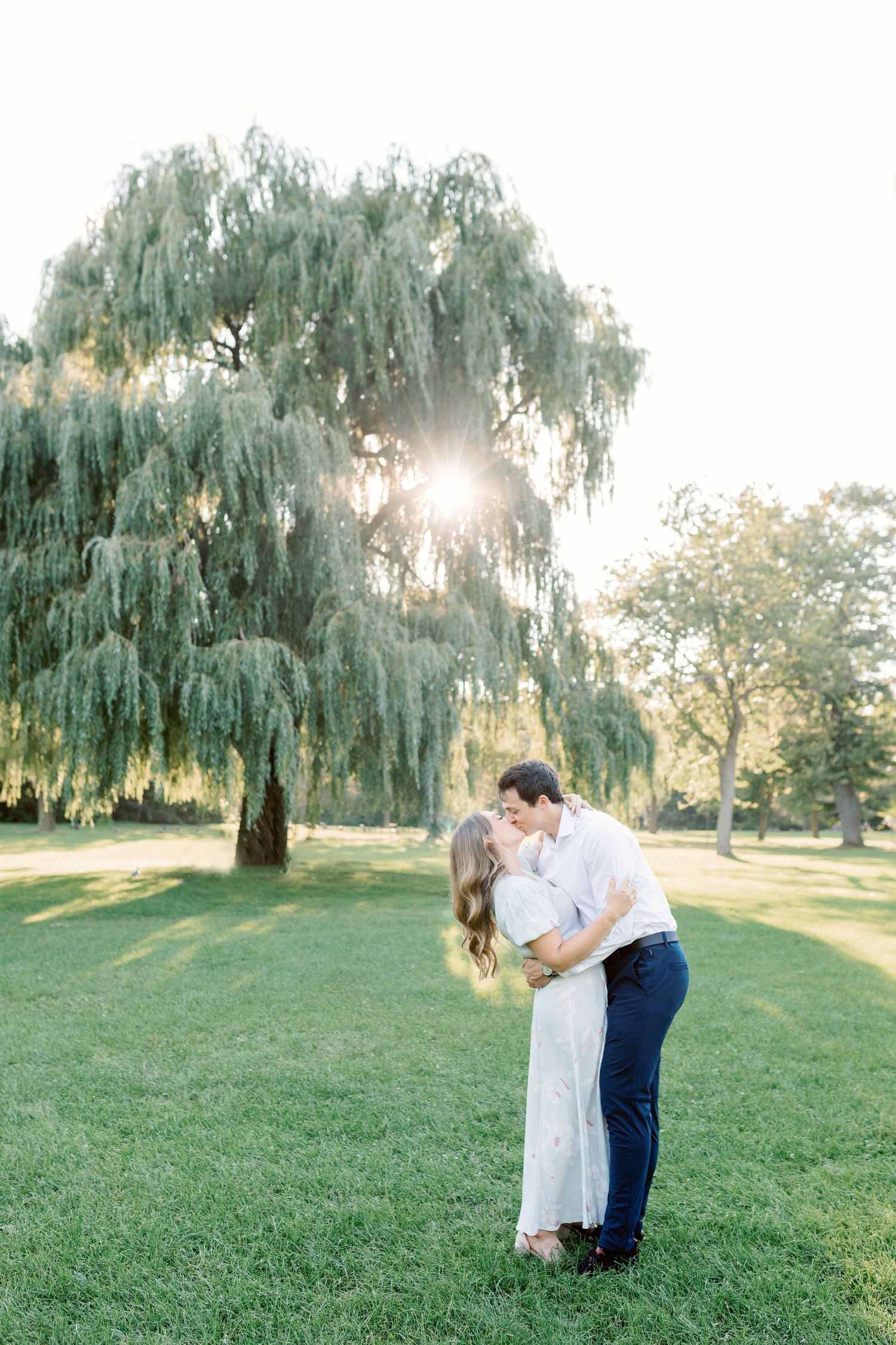 A romantic couple embraces under a weeping willow tree at sunset in a park.