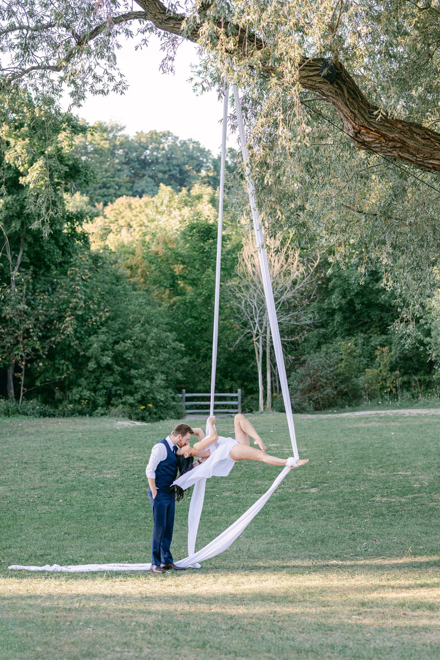 Couple playing on white fabric swings hanging from a tree branch in a grassy park setting.