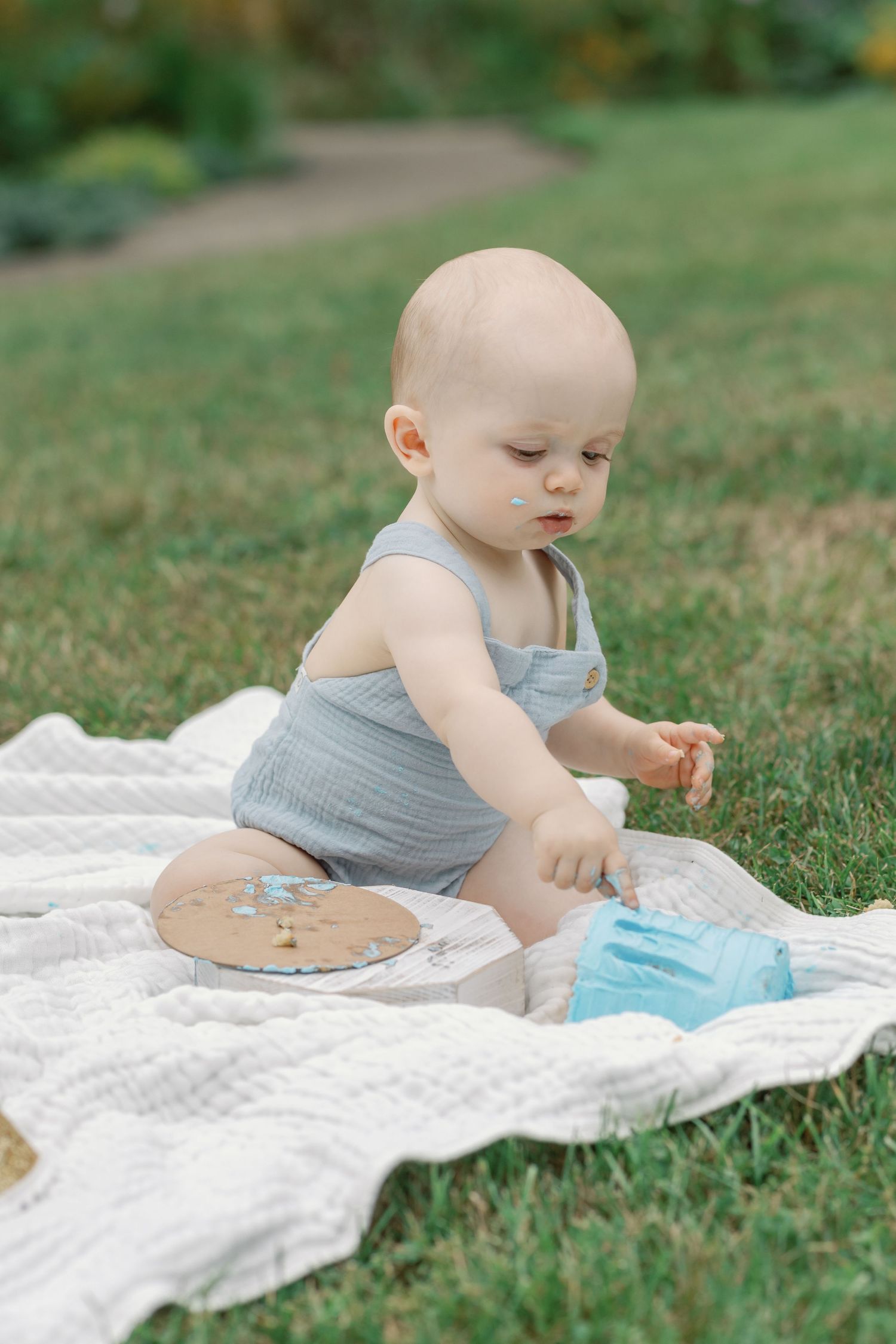 Baby in sleeveless gray outfit explores toys while sitting on white blanket outdoors.