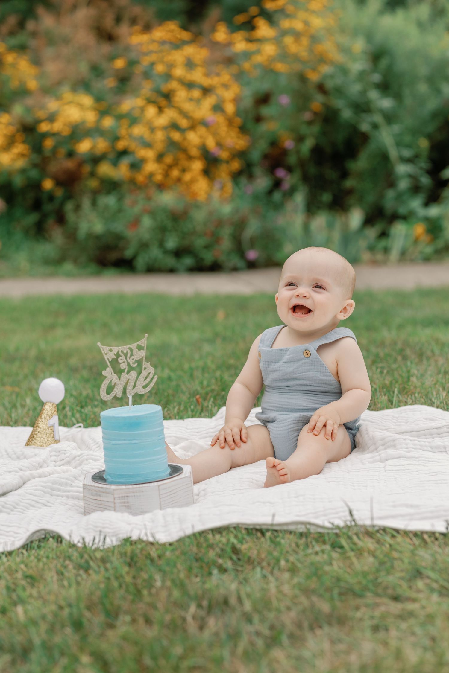 Smiling baby explores first birthday setup with blue cake on white blanket outdoors.