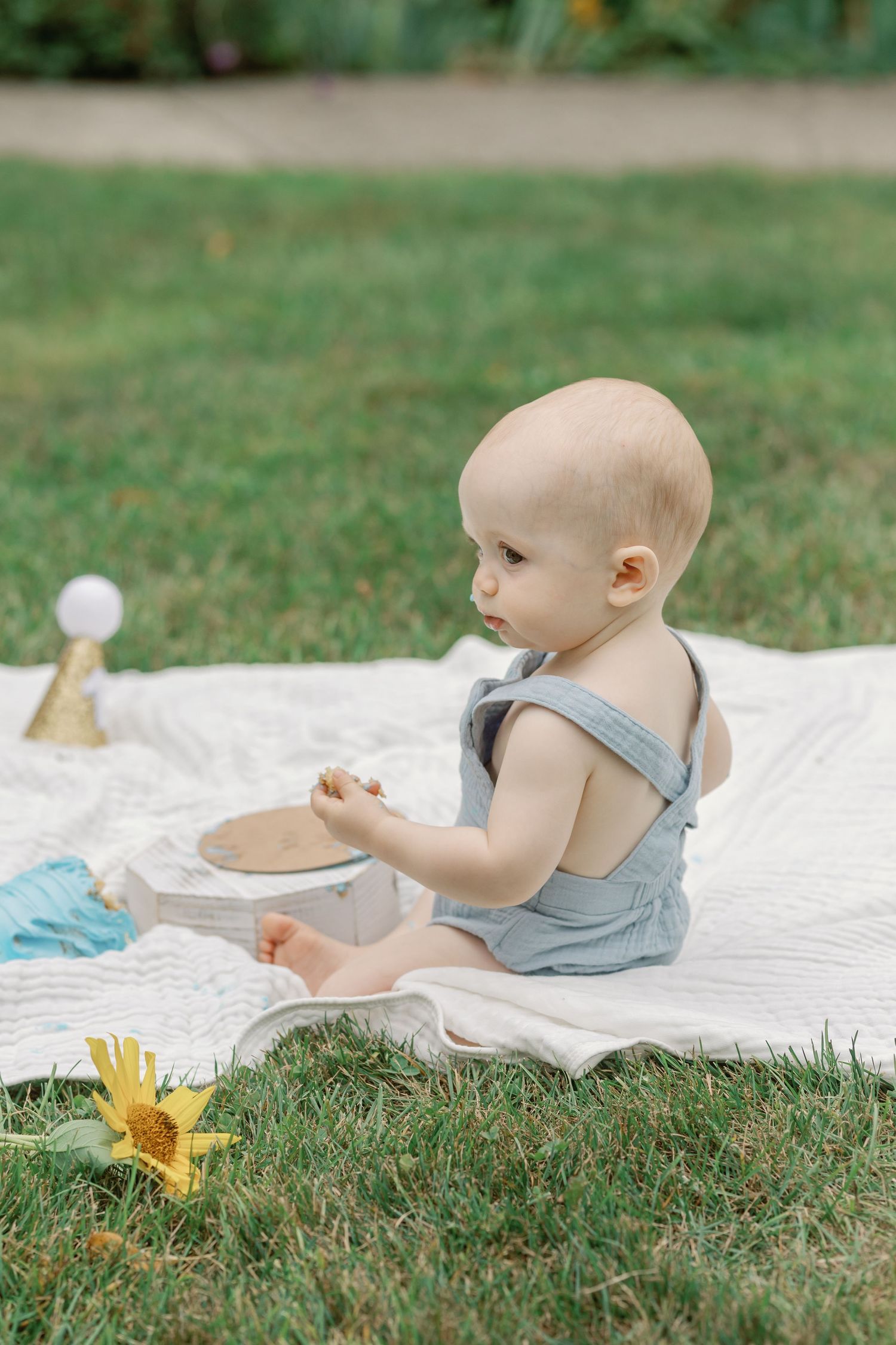 Baby in gray outfit plays with toys on white blanket during outdoor photoshoot.