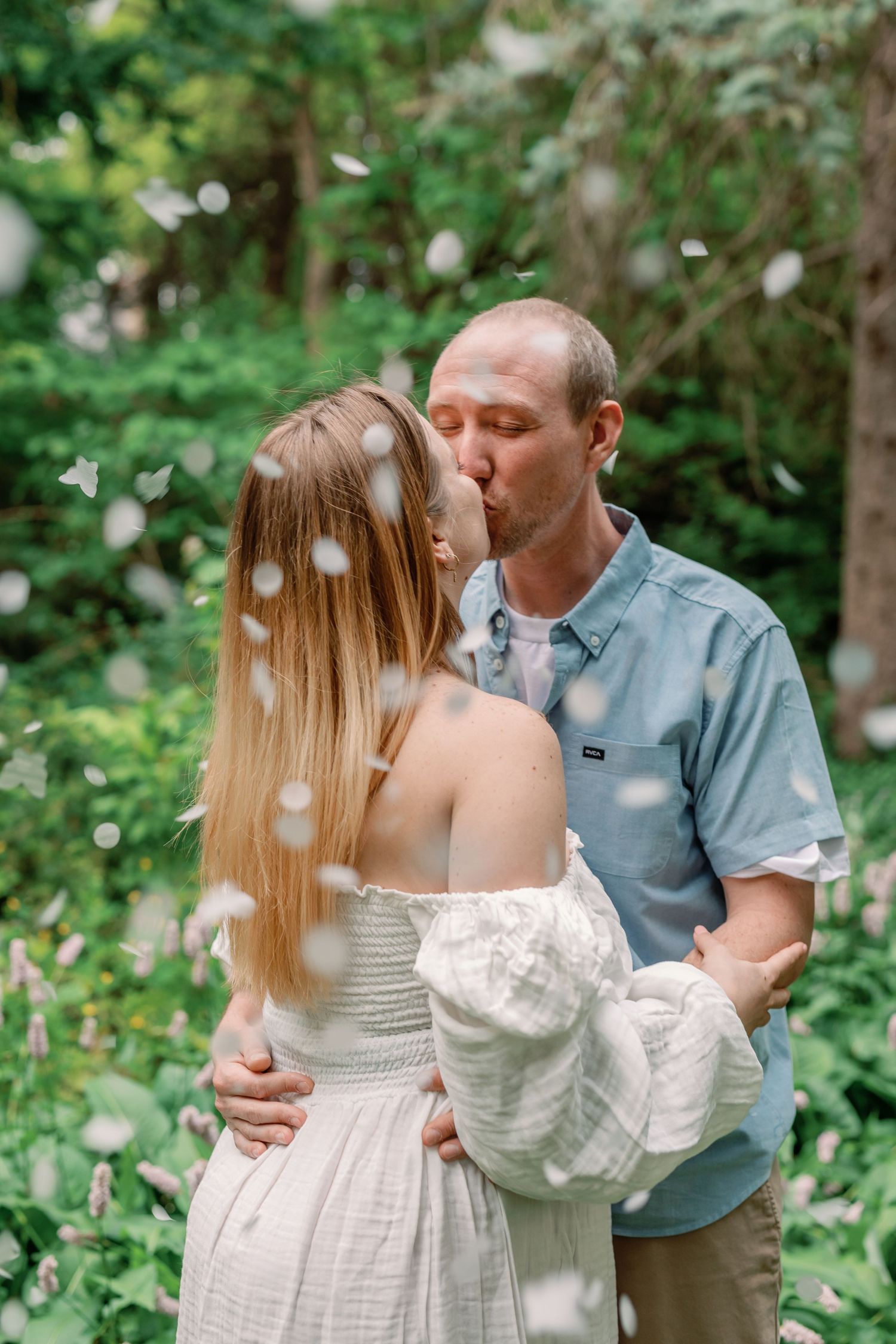 Couple embraces outdoors surrounded by greenery and natural light.