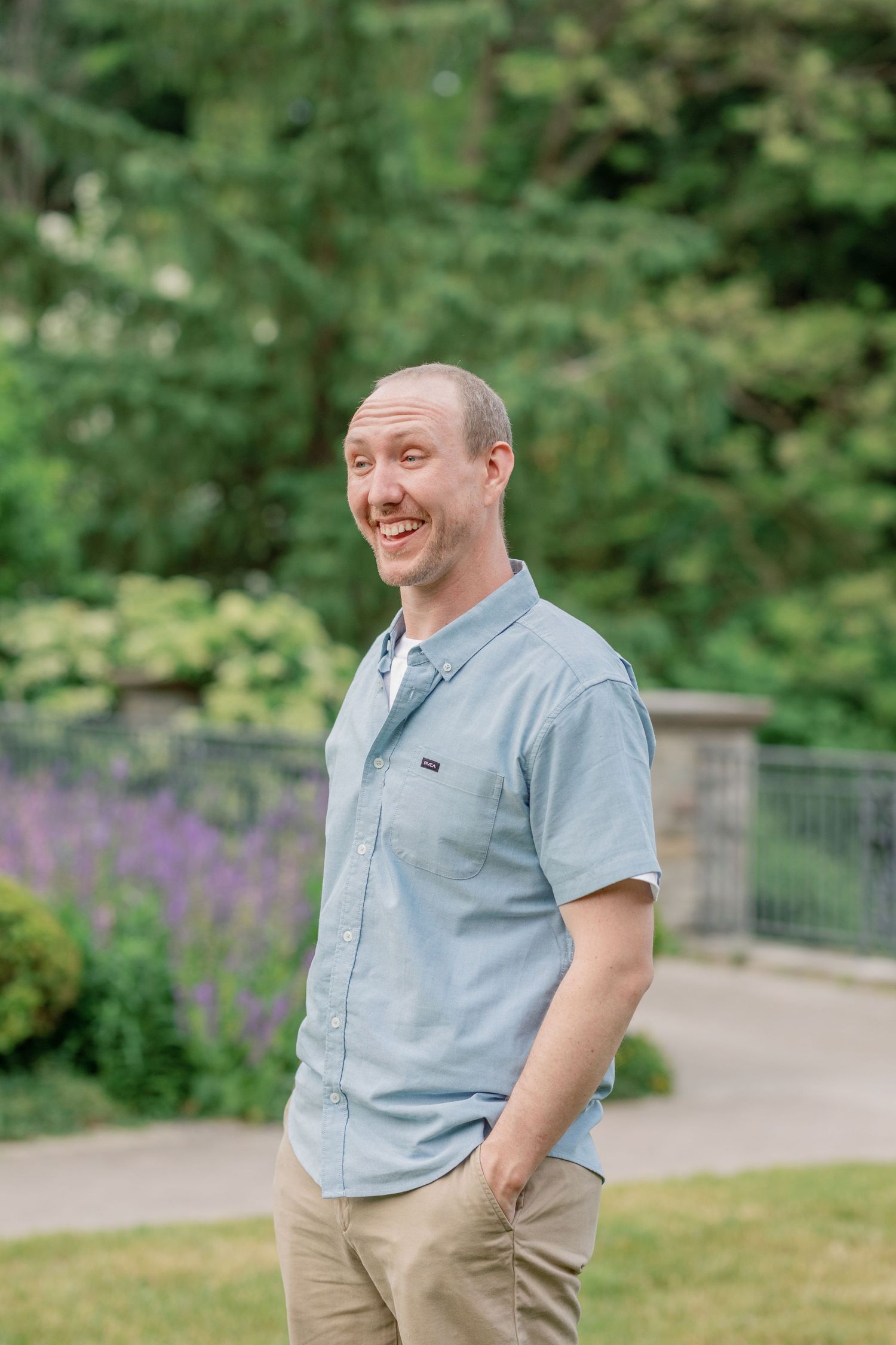 A person in a light blue shirt smiles warmly while standing in a garden with purple flowers blooming.