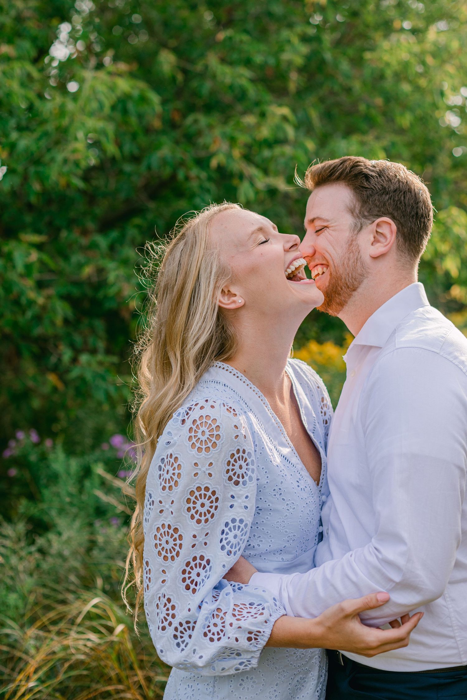A joyful couple shares laughter in a garden setting wearing white and light colored clothing.