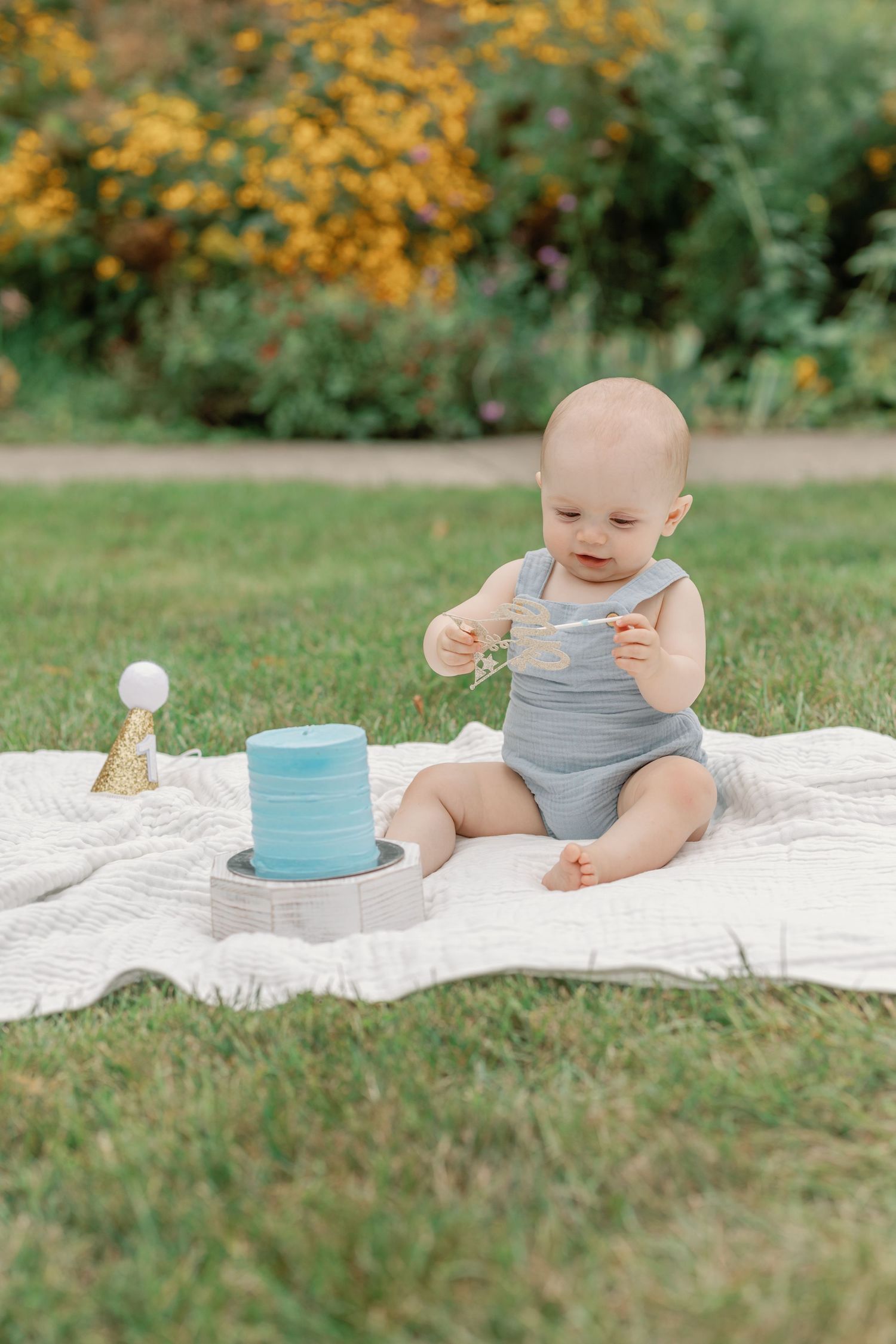 Baby in grey romper sits on white blanket outdoors with blue birthday cake and decorations.