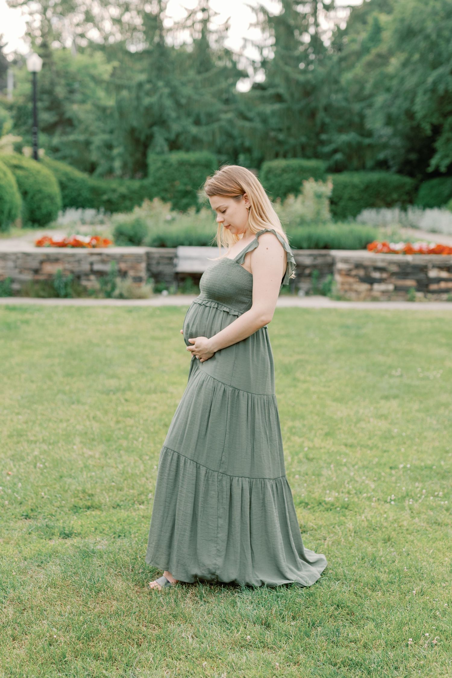 Person in a sage green dress standing in a garden with stone walls and evergreen trees.