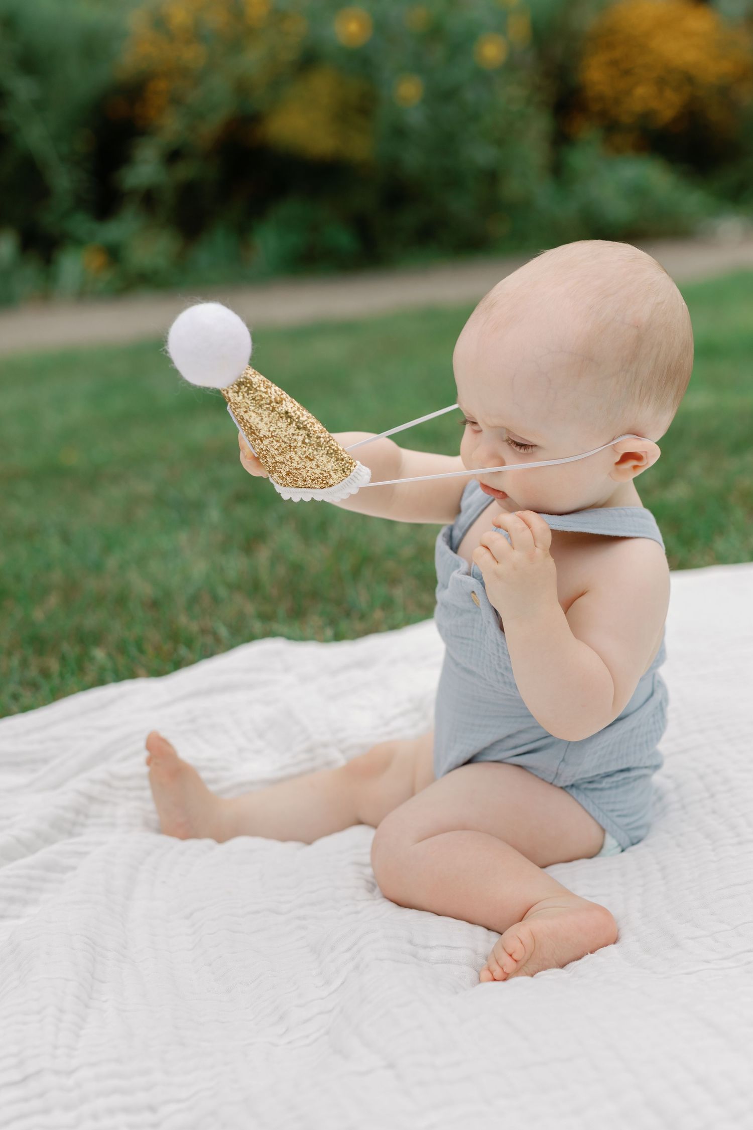 Baby in grey outfit plays with birthday decorations on white blanket in garden.