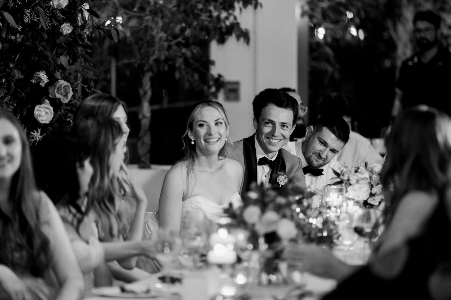 A bride and groom laugh with their guests at the head table, decorated with flower bouquets. Photo: Katie Crane Photogra