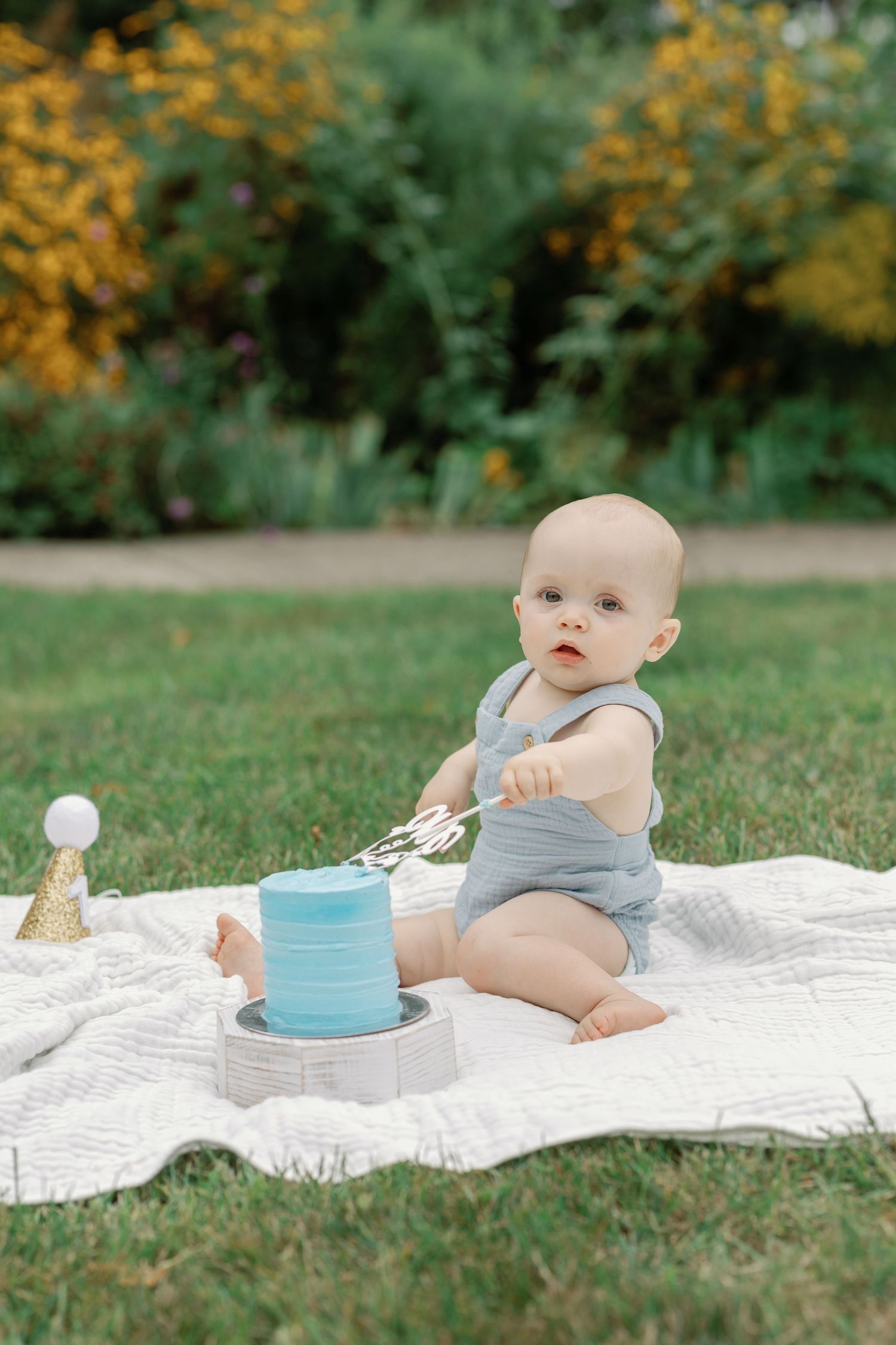 Baby in gray romper sits on white blanket with yellow flowers in background.
