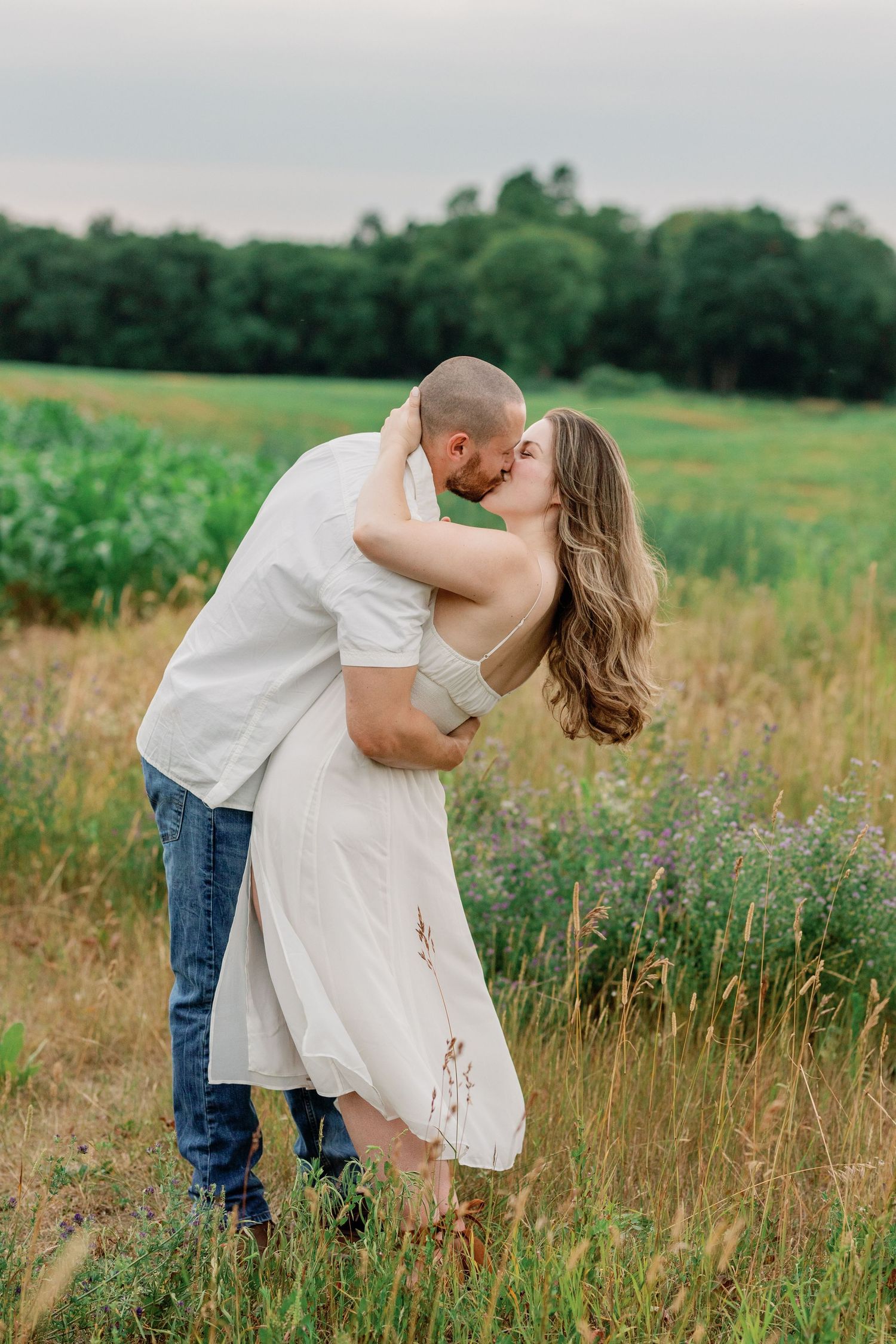 A romantic embrace in a sunlit meadow with tall grass and trees in the background.