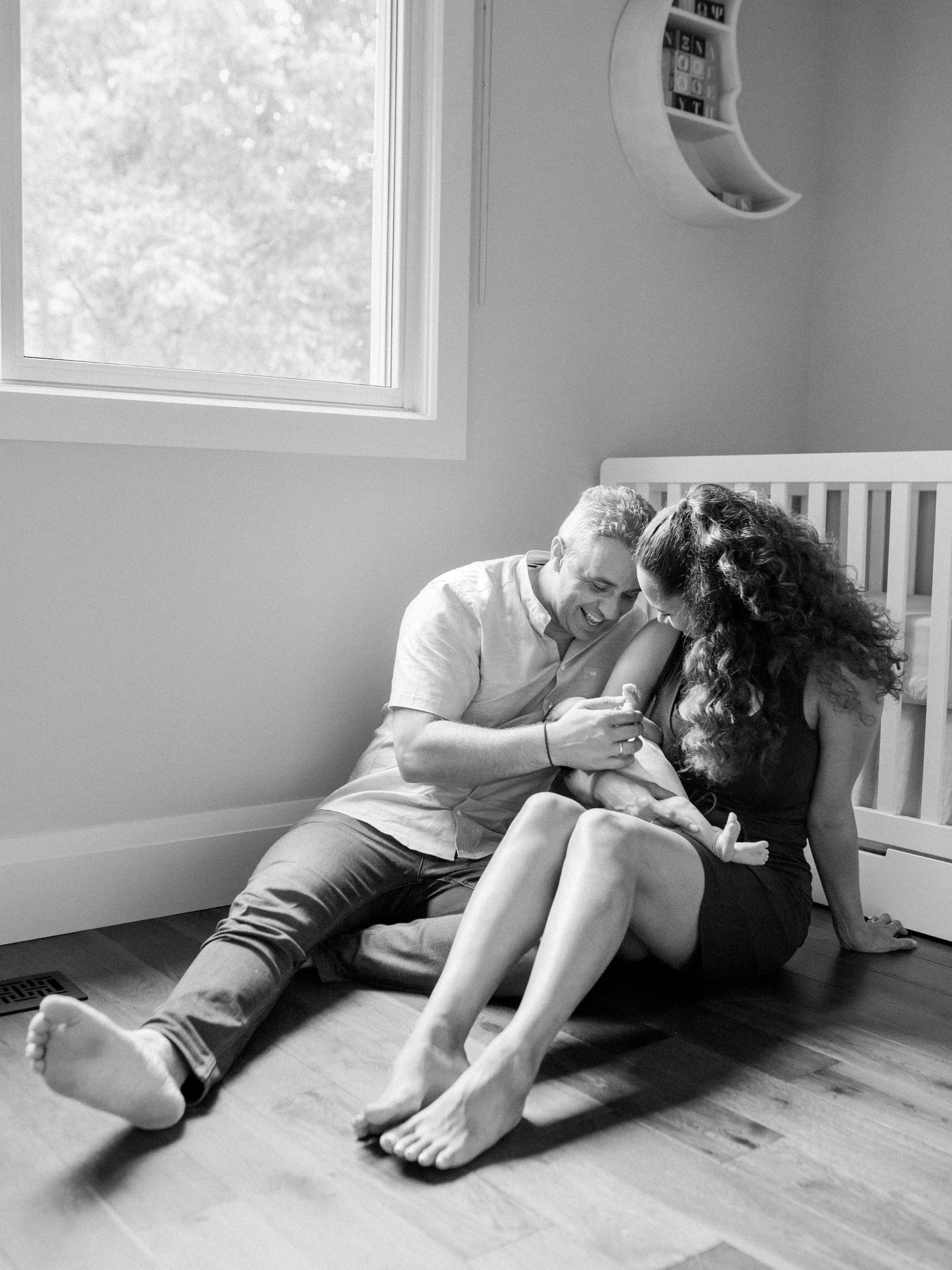 A mother and father sit together against the cradle and smile down at their newborn in their arms. Photo of Katie Crane 