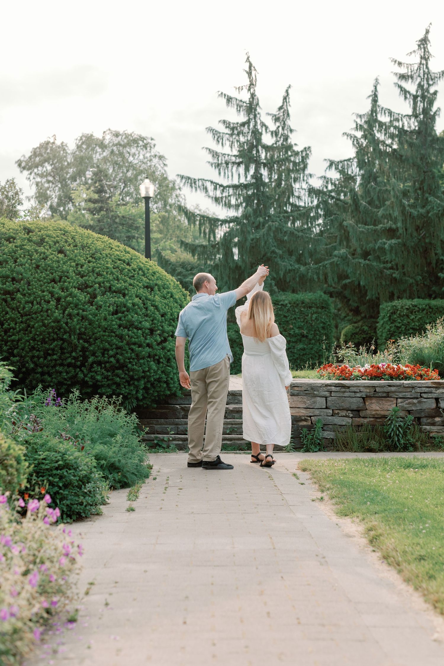 A couple walks together on a garden path surrounded by evergreen trees and colorful flowers.