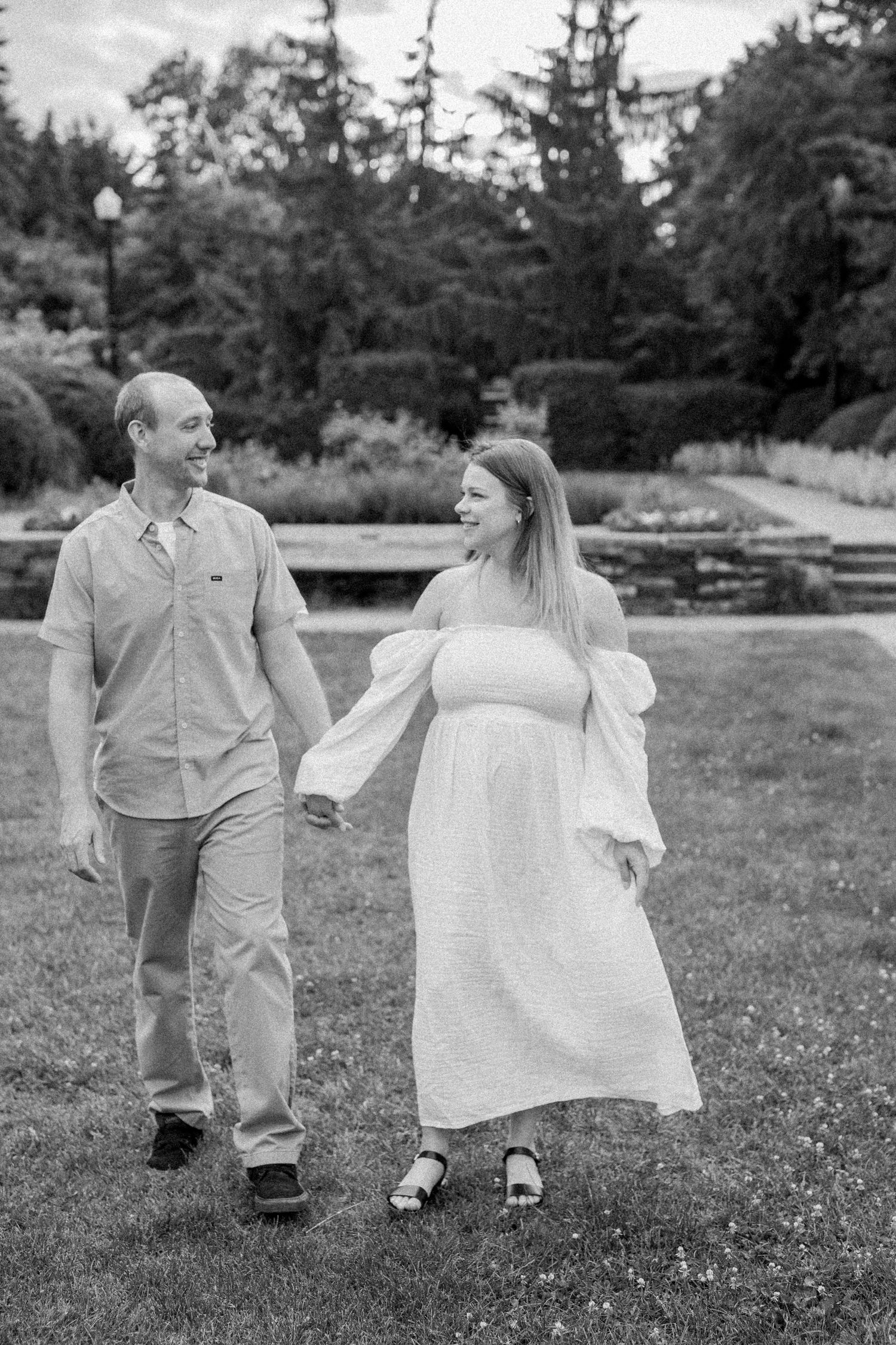 A couple in light colored clothing walks playfully through a grassy park area.