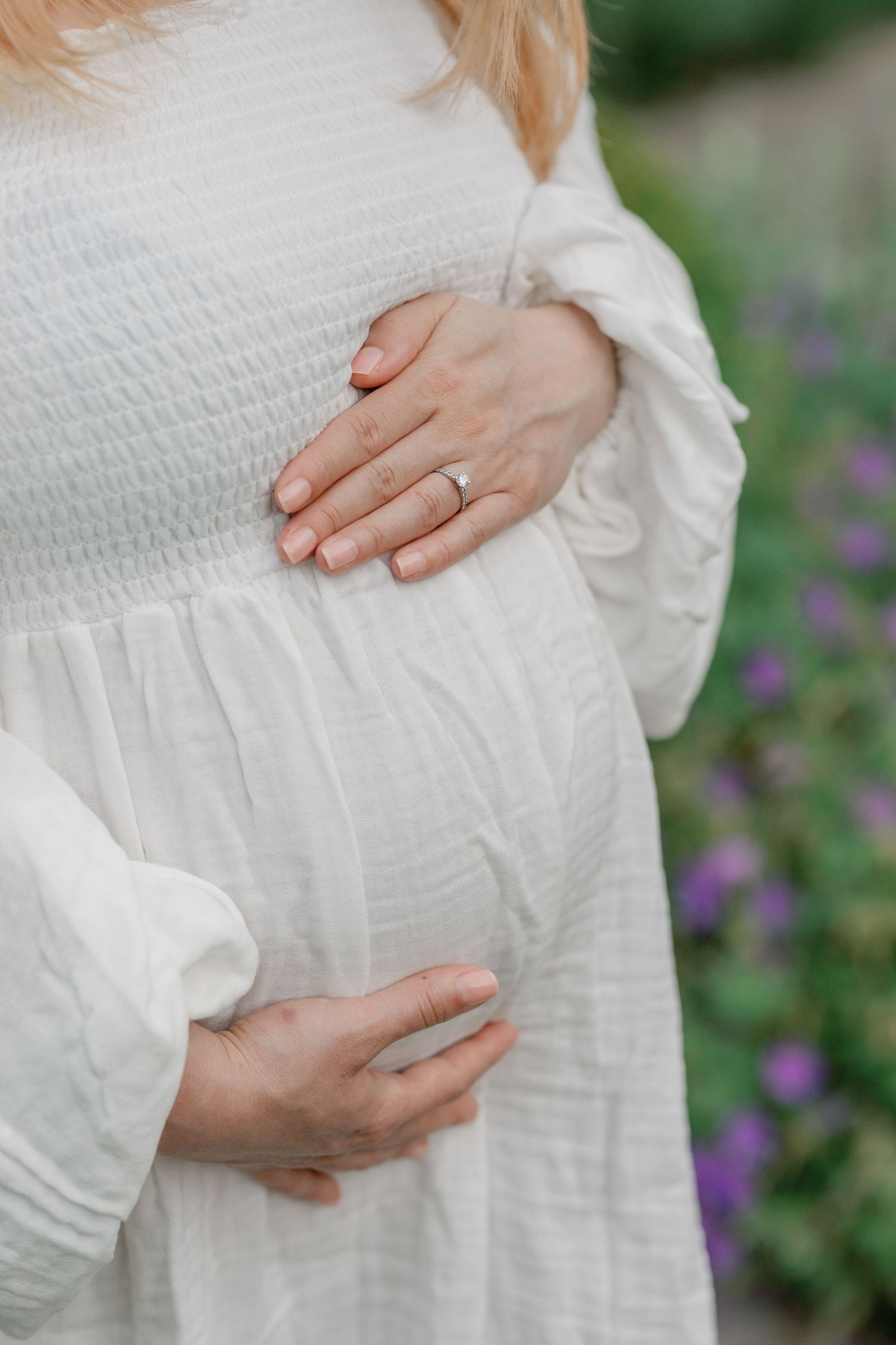 Close-up of hands cradling a pregnant belly in a white dress against purple flowers in the background.