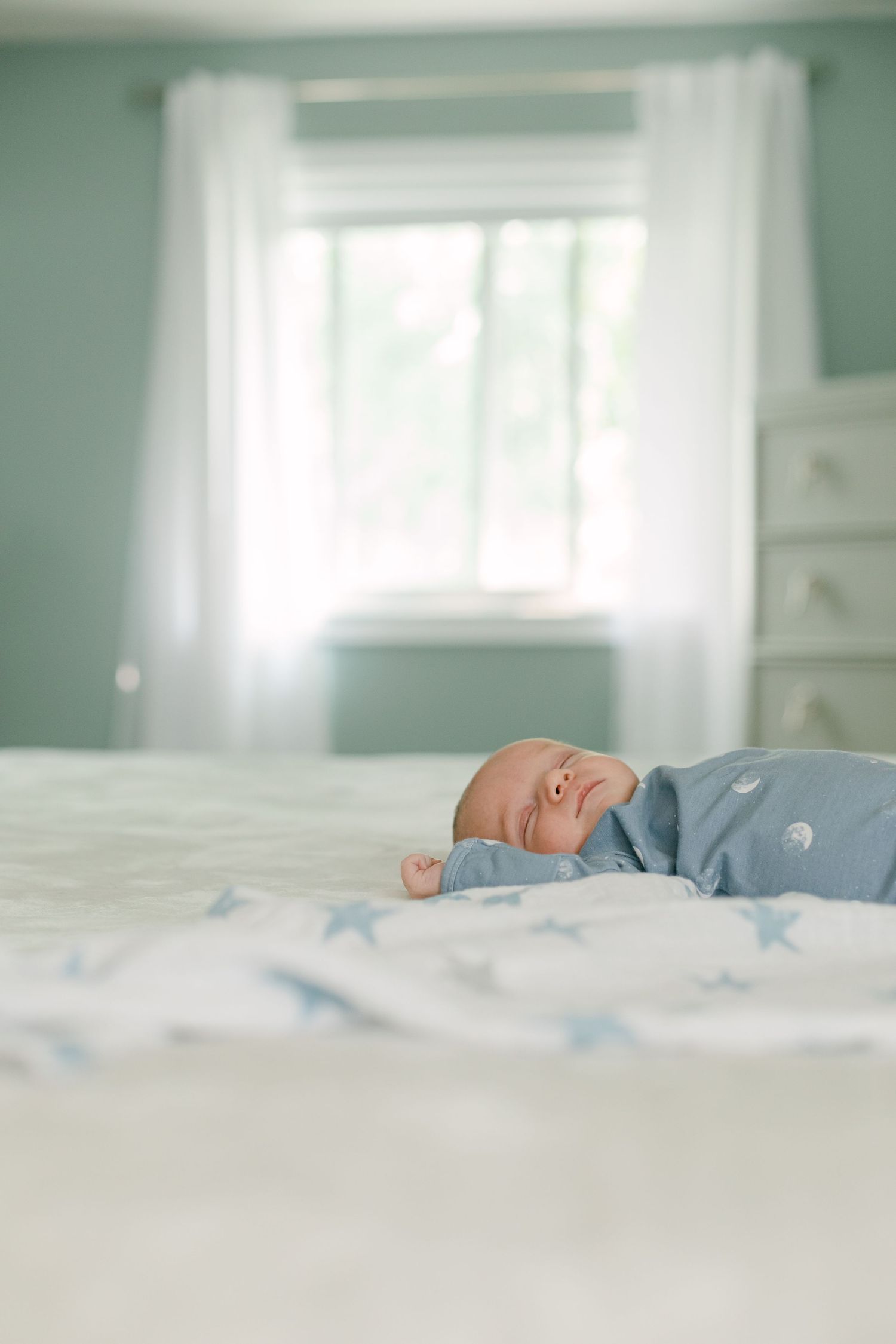 A baby in a blue jumper lies sleeping on a bed next to a blanket covered in blue stars, the walls of the room are blue. 