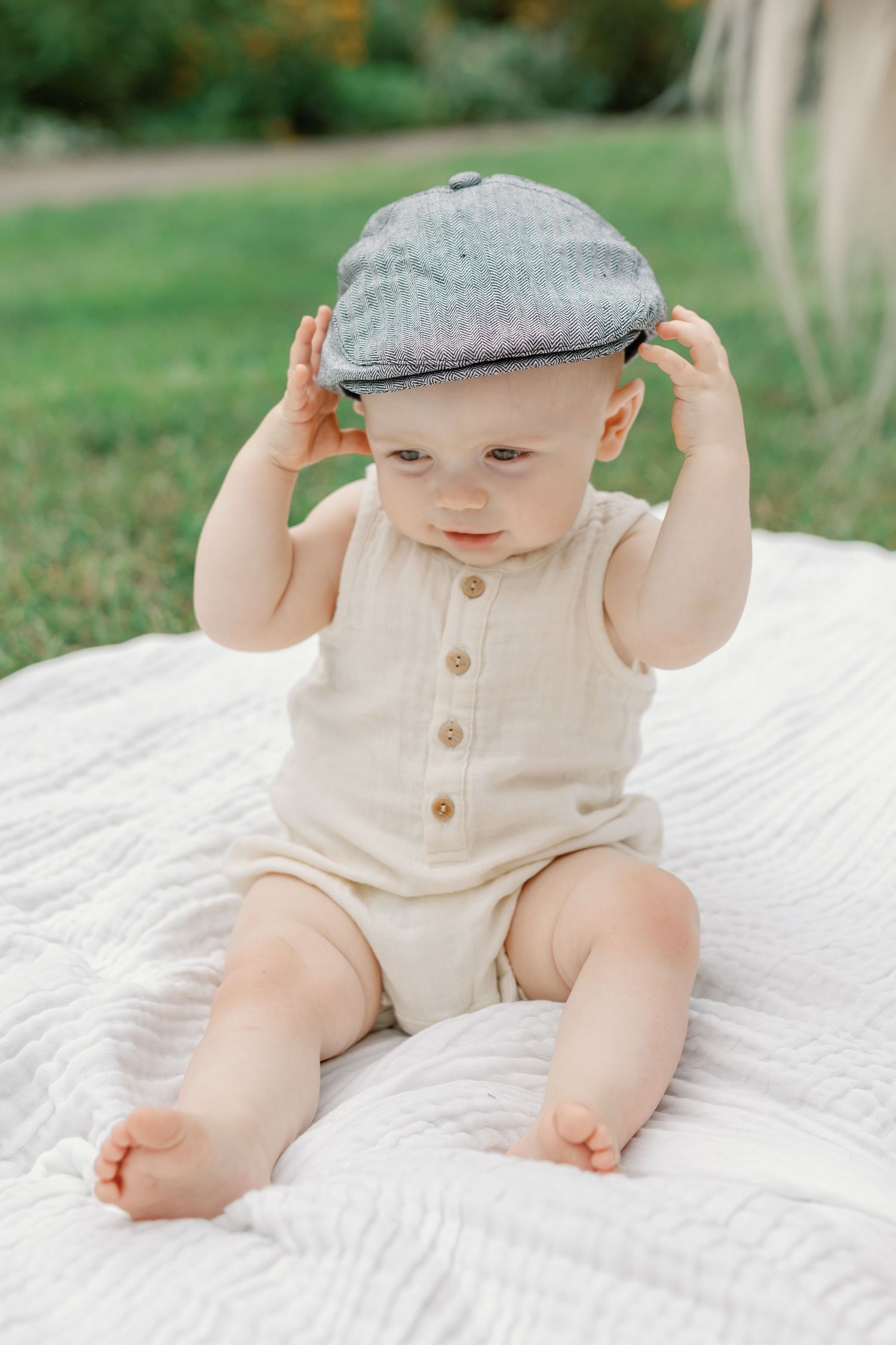 Baby wearing newsboy cap and cream romper sits on white blanket outdoors.