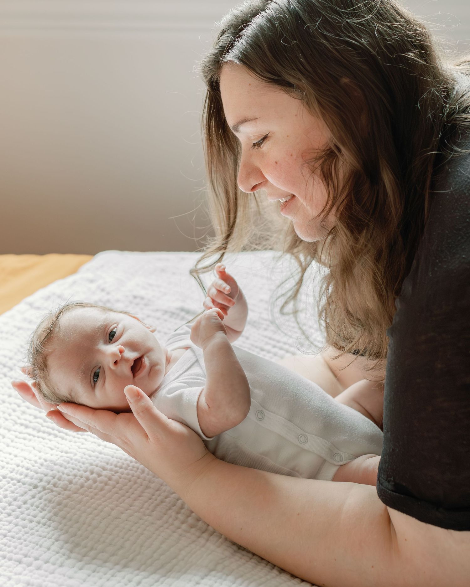 A mother holds her newborn in one arm, laying him down on the yellow sheets of a bed. Photo by Katie Crane.