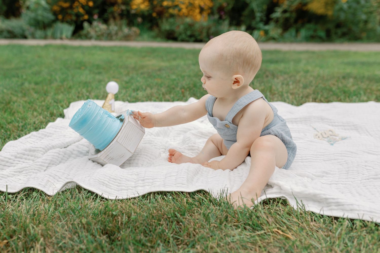 Baby in gray outfit plays on white blanket during outdoor summer photoshoot.