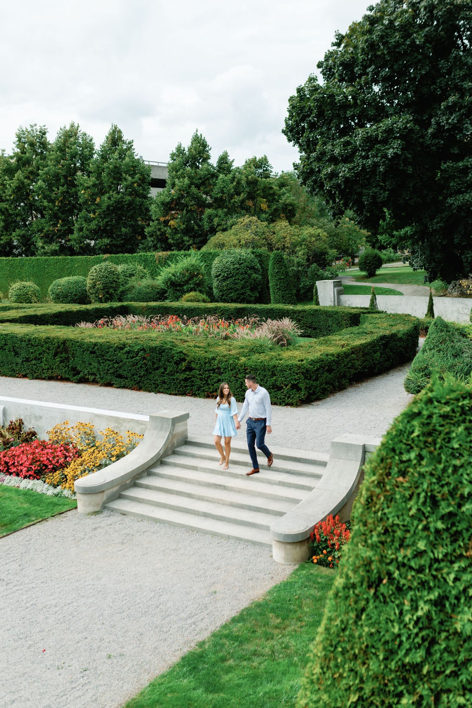 Couple walking down garden steps surrounded by manicured hedges and colorful flowers.