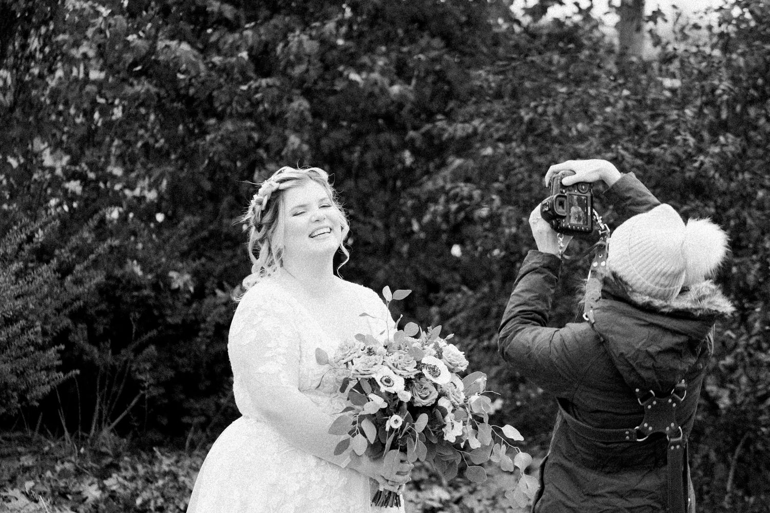 Katie takes a photo of a bride in her dress standing in front of greenery.