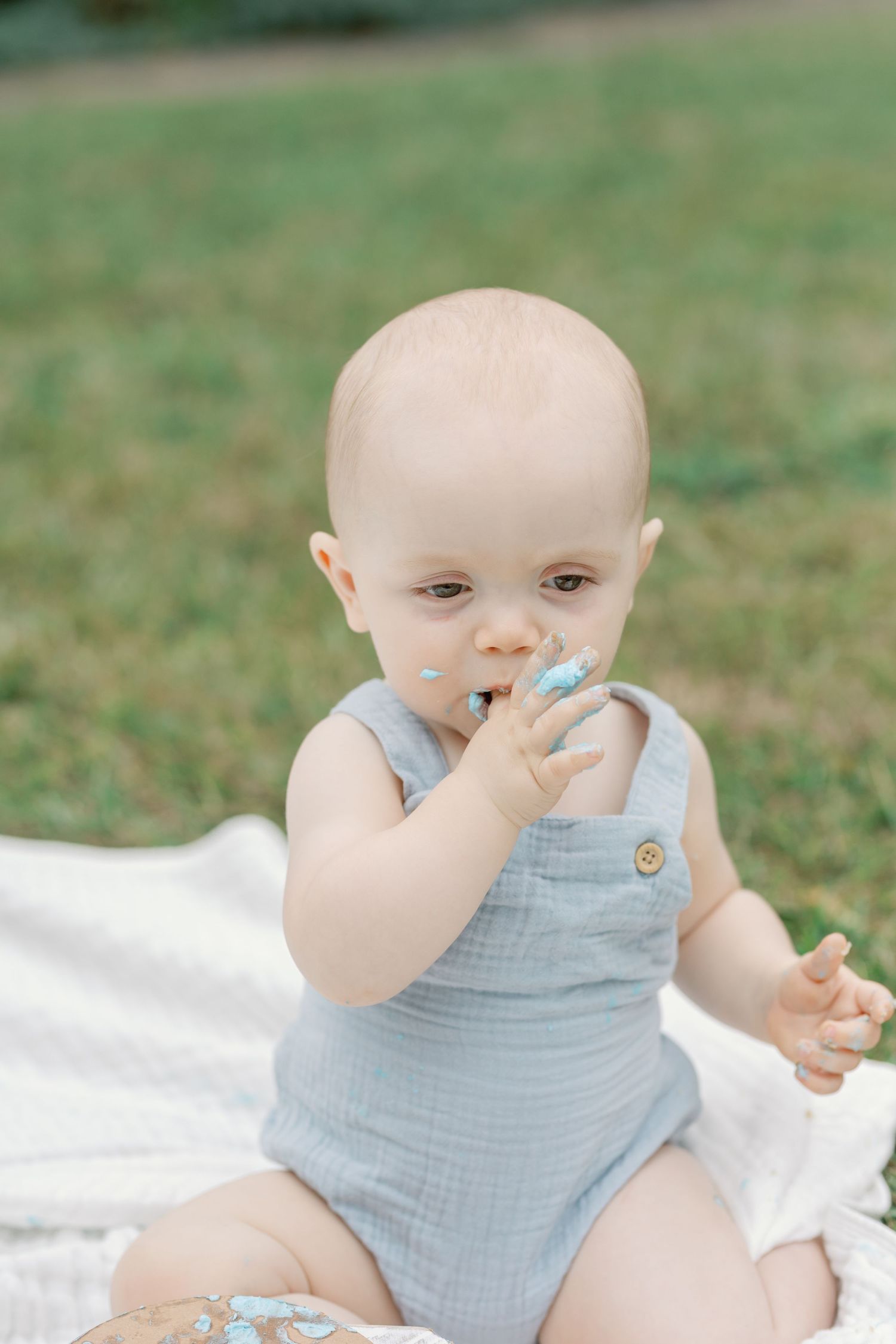 Baby wearing blue romper plays on white blanket during outdoor photo session.