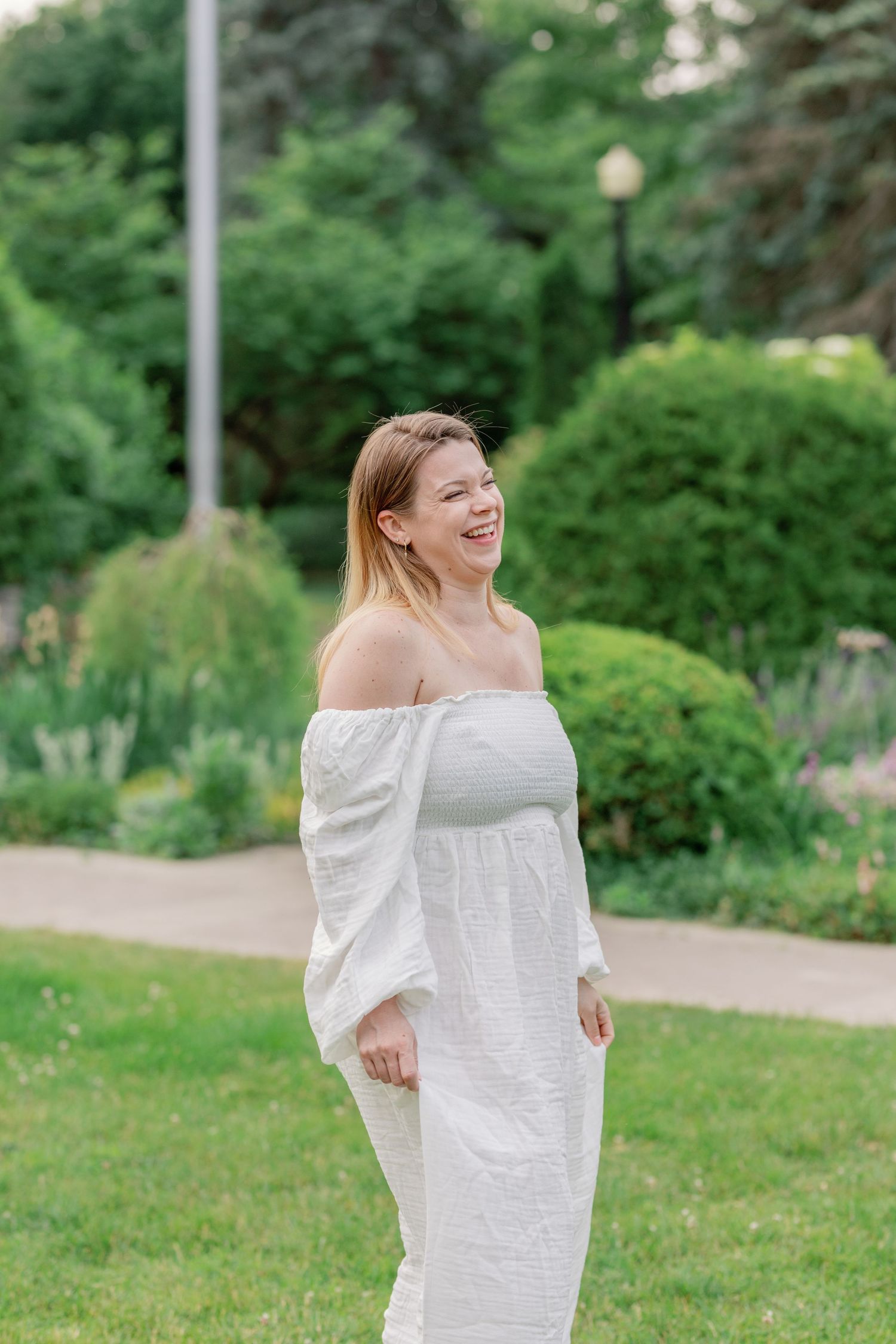 A woman in a white off-shoulder dress poses playfully in a garden setting with green foliage.