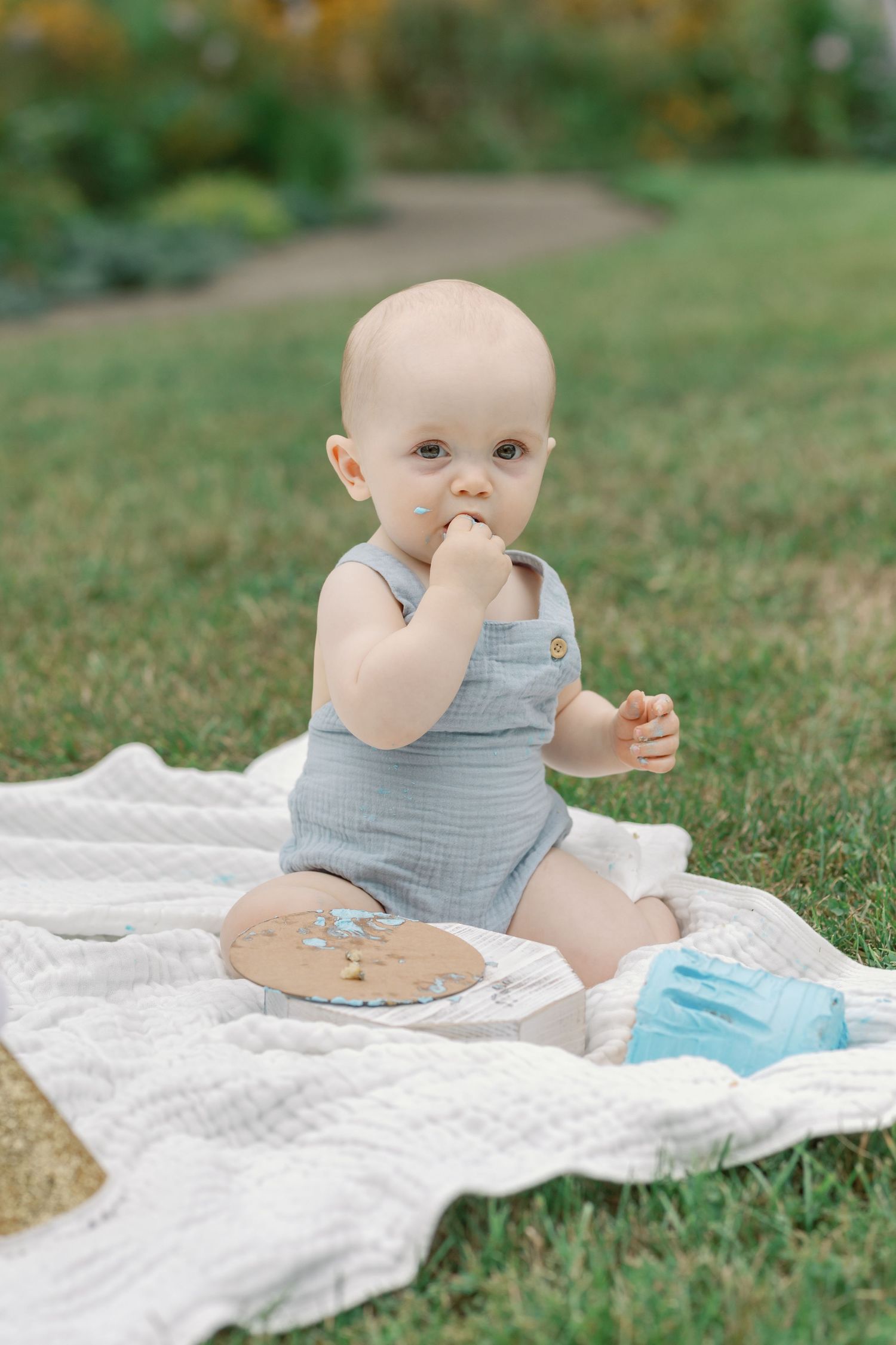 Baby in gray outfit sits on white blanket during outdoor photography session.