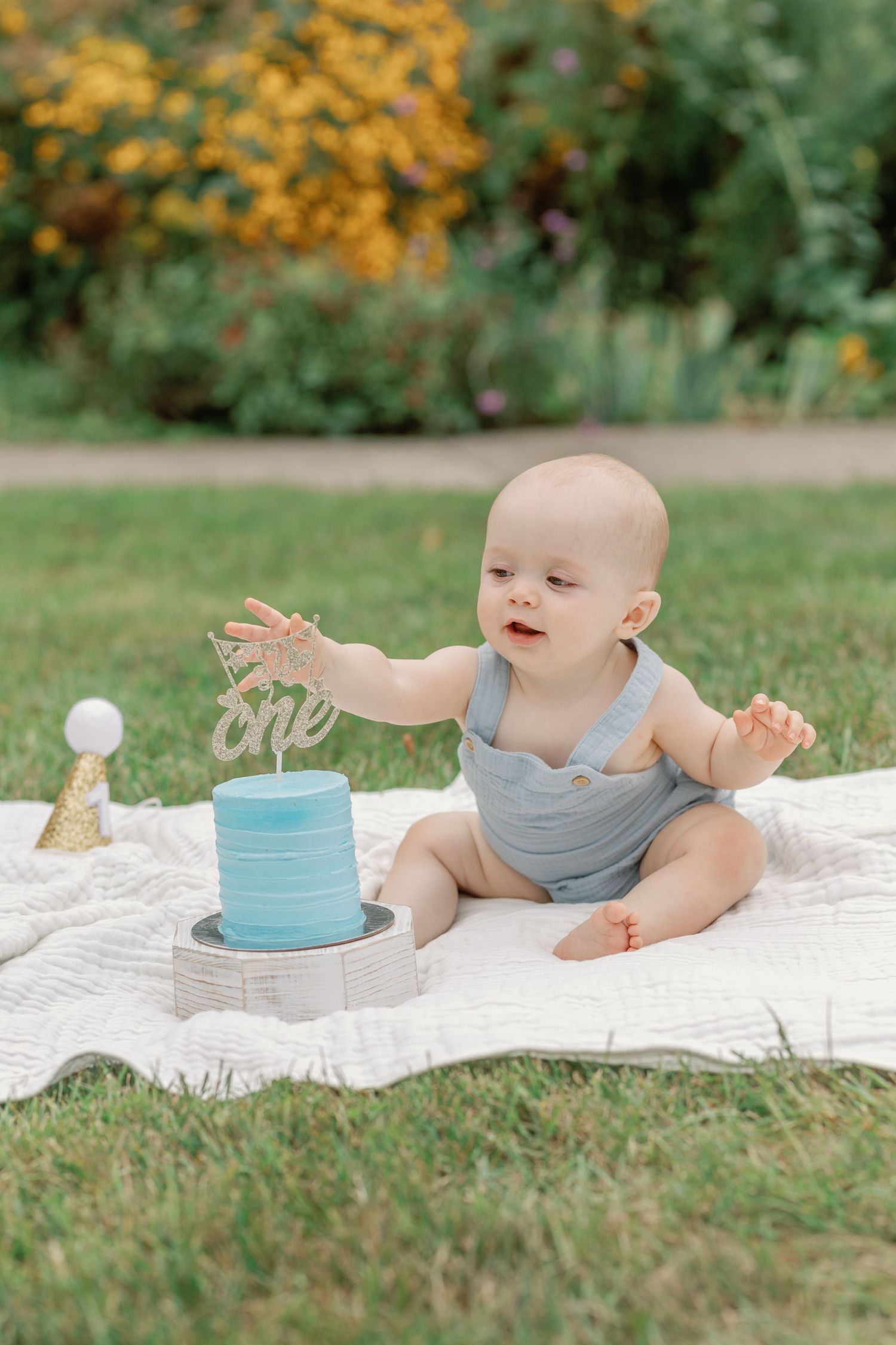 Baby reaches towards blue cake while sitting on white blanket in garden setting.
