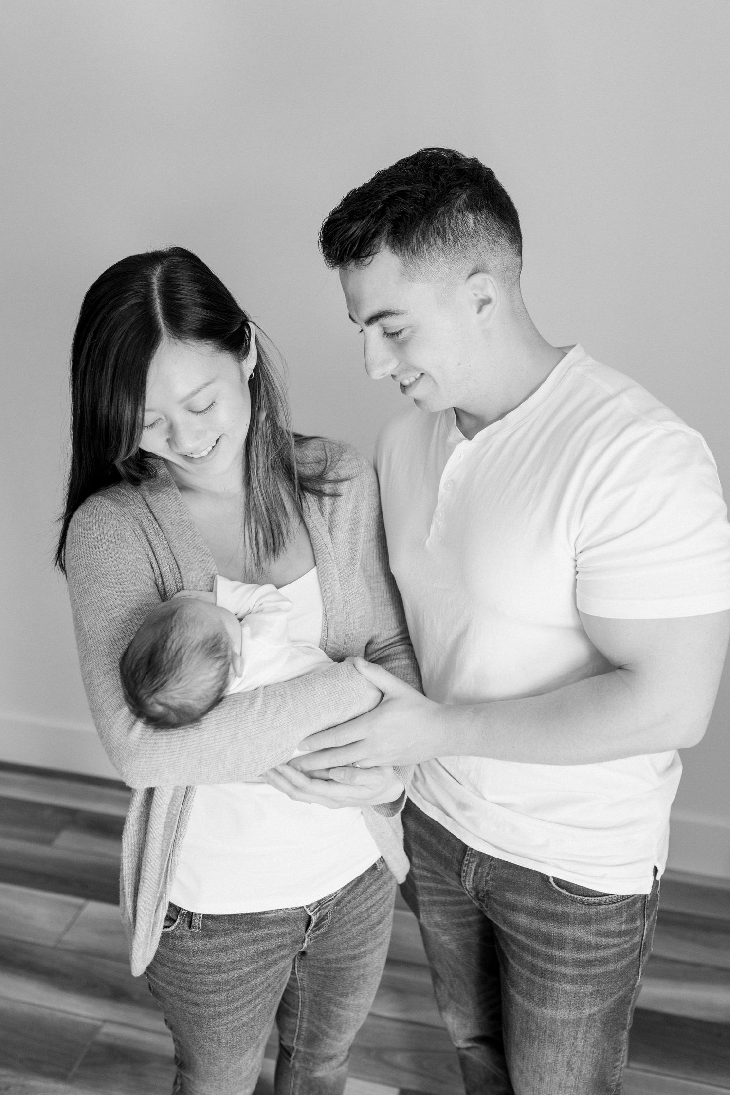 A young couple stands lookingl lovingly at their newborn who is held by the mother. Photo by Katie Crane Photography.