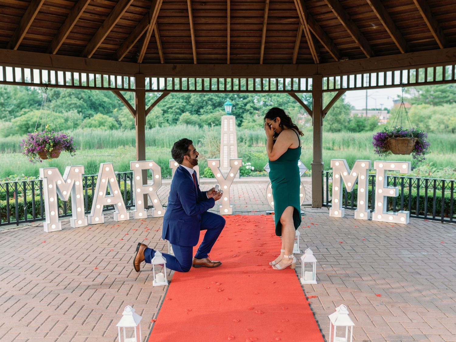 A marriage proposal scene in a gazebo with red carpet and illuminated 'MARRY ME' letters.