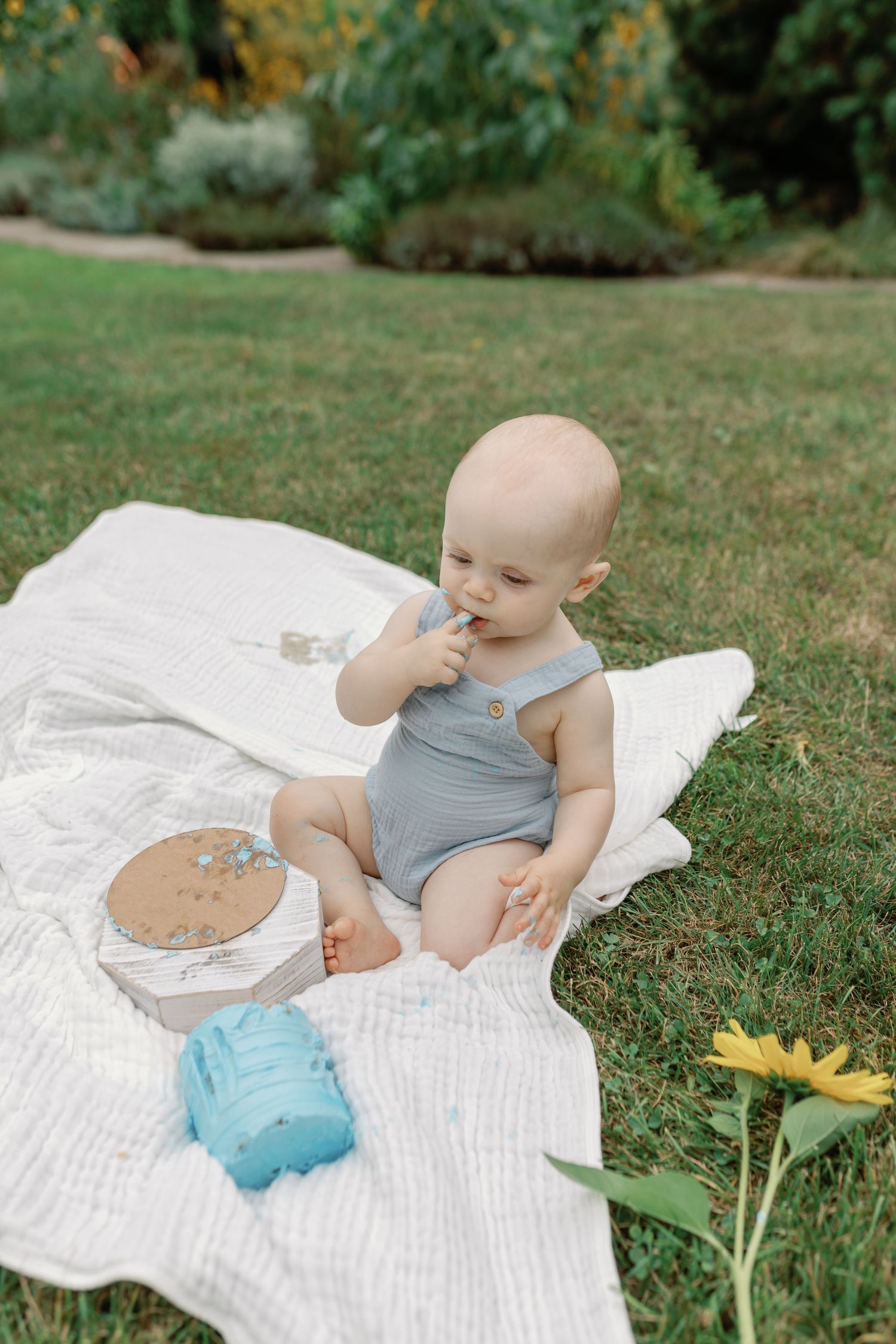 Baby in blue romper explores toys on white blanket with yellow flower in background.