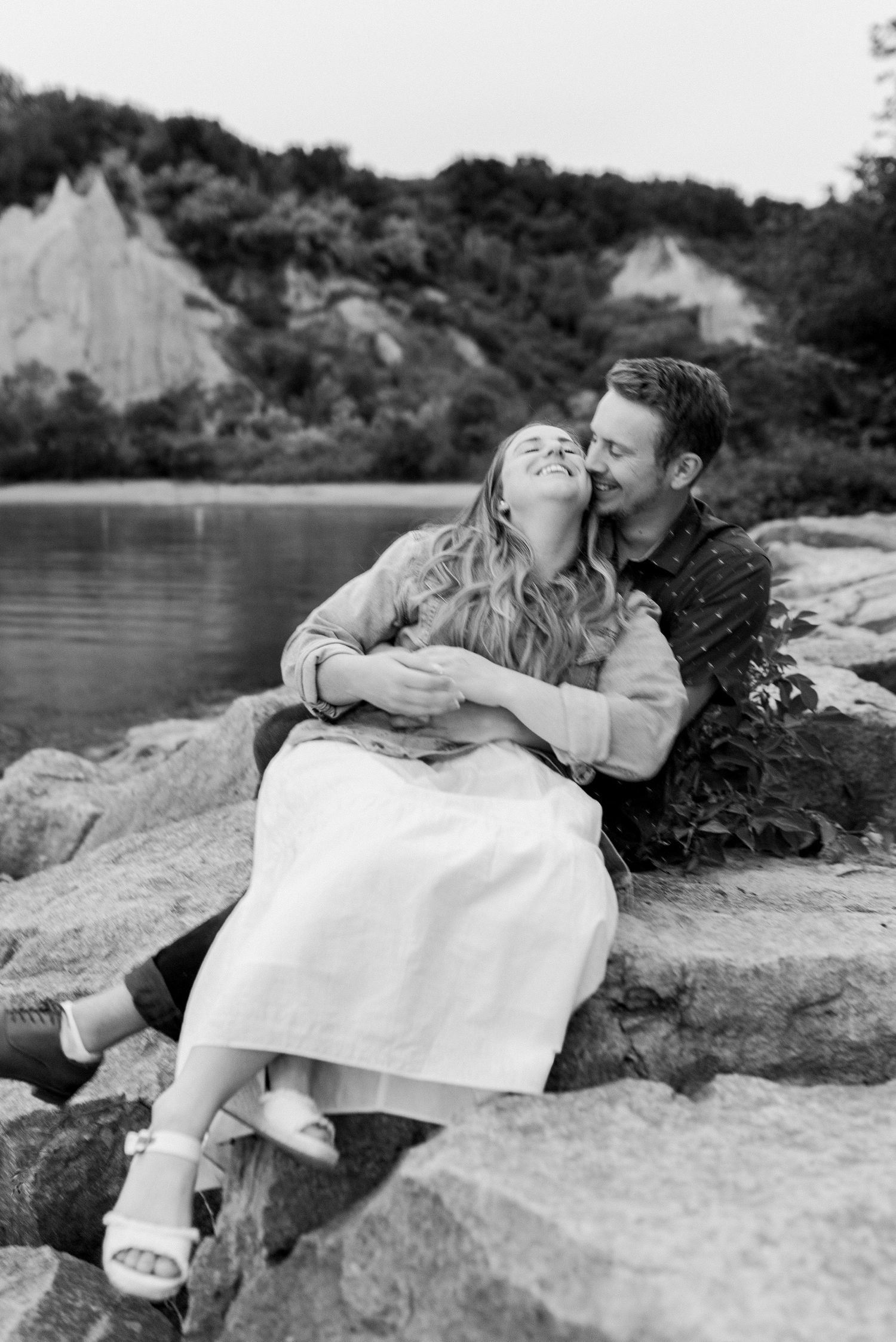 A black and white photo of a couple sharing an intimate moment while sitting on rocky cliffs by a lake.