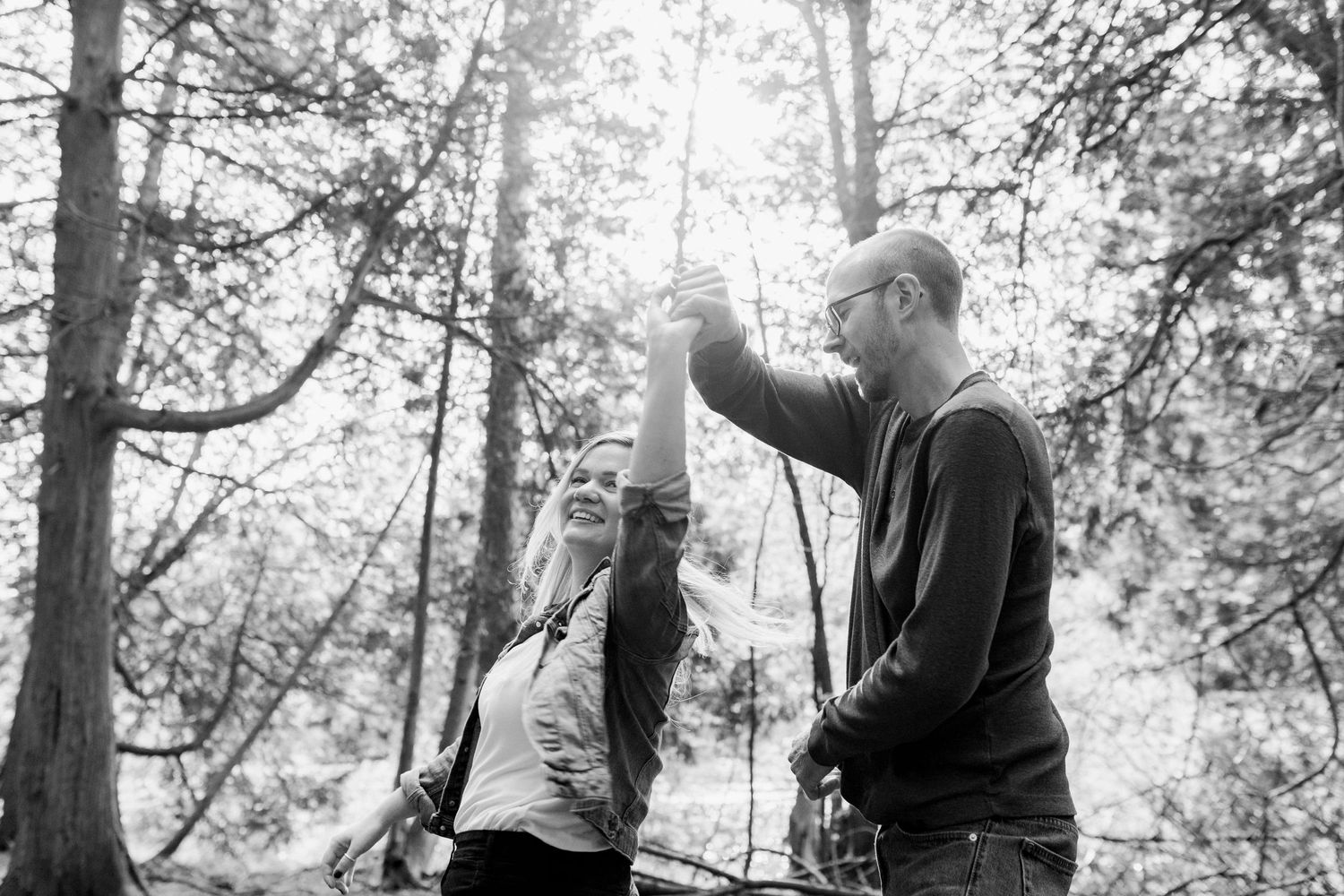 A playful moment captured in black and white as a couple dances in a sunlit forest.