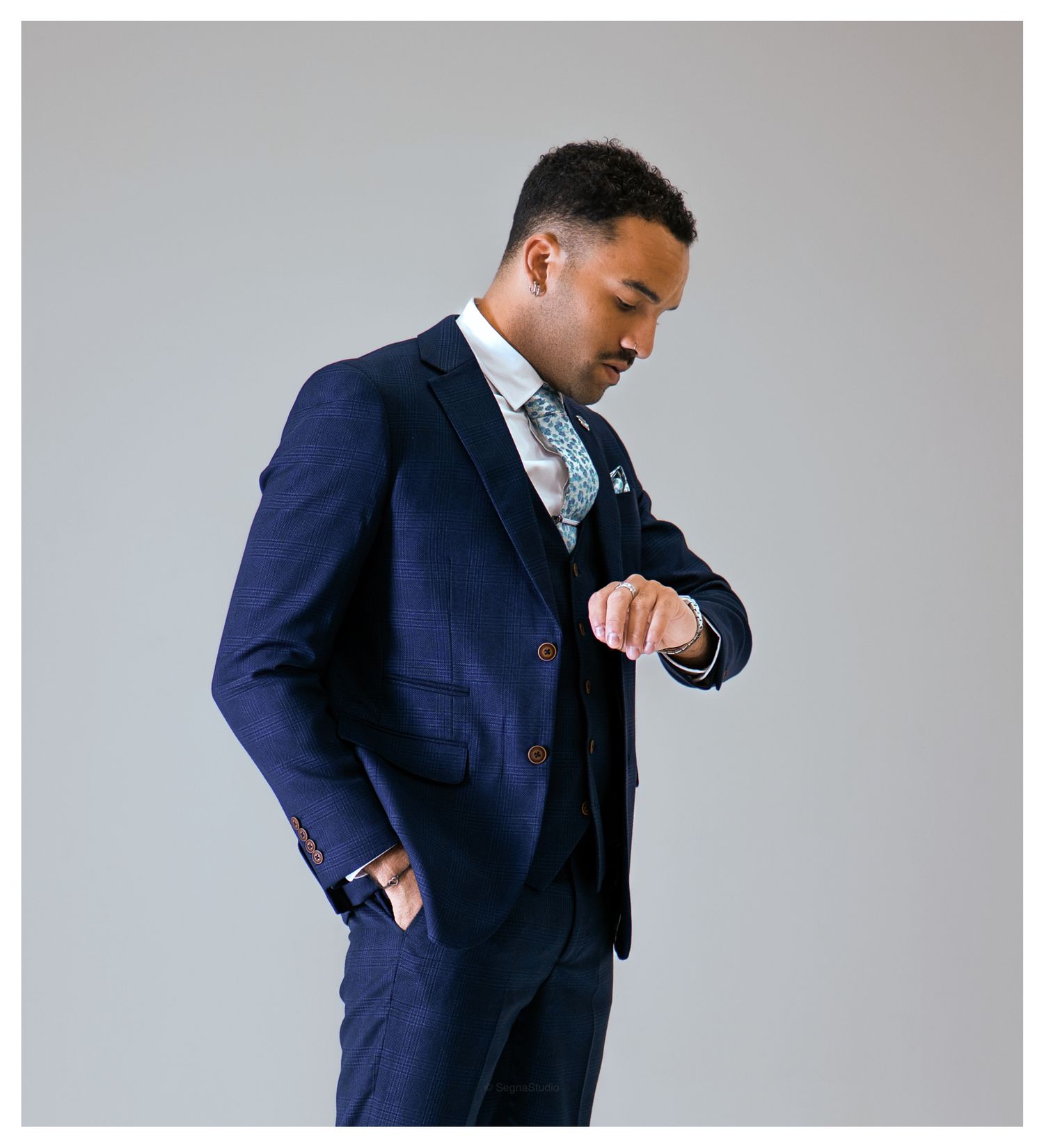 Man in a navy three-piece suit looking at his wrist against a neutral grey background.