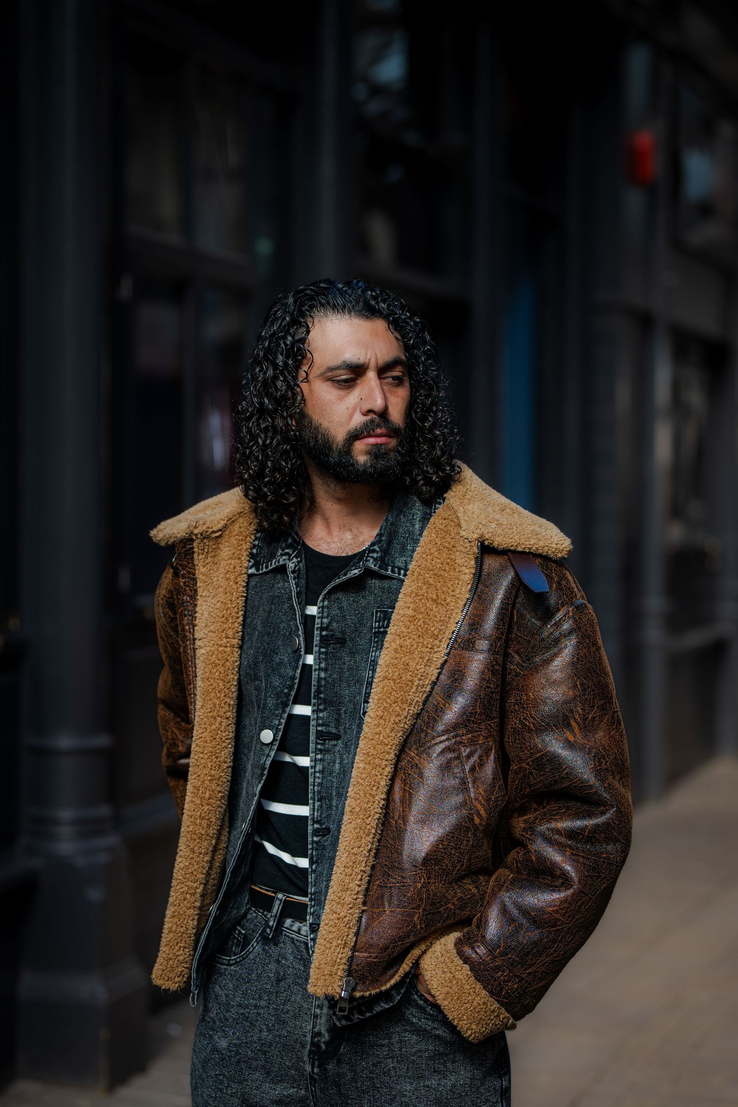 Man with curly hair and beard wearing a brown shearling leather jacket standing on an urban street.