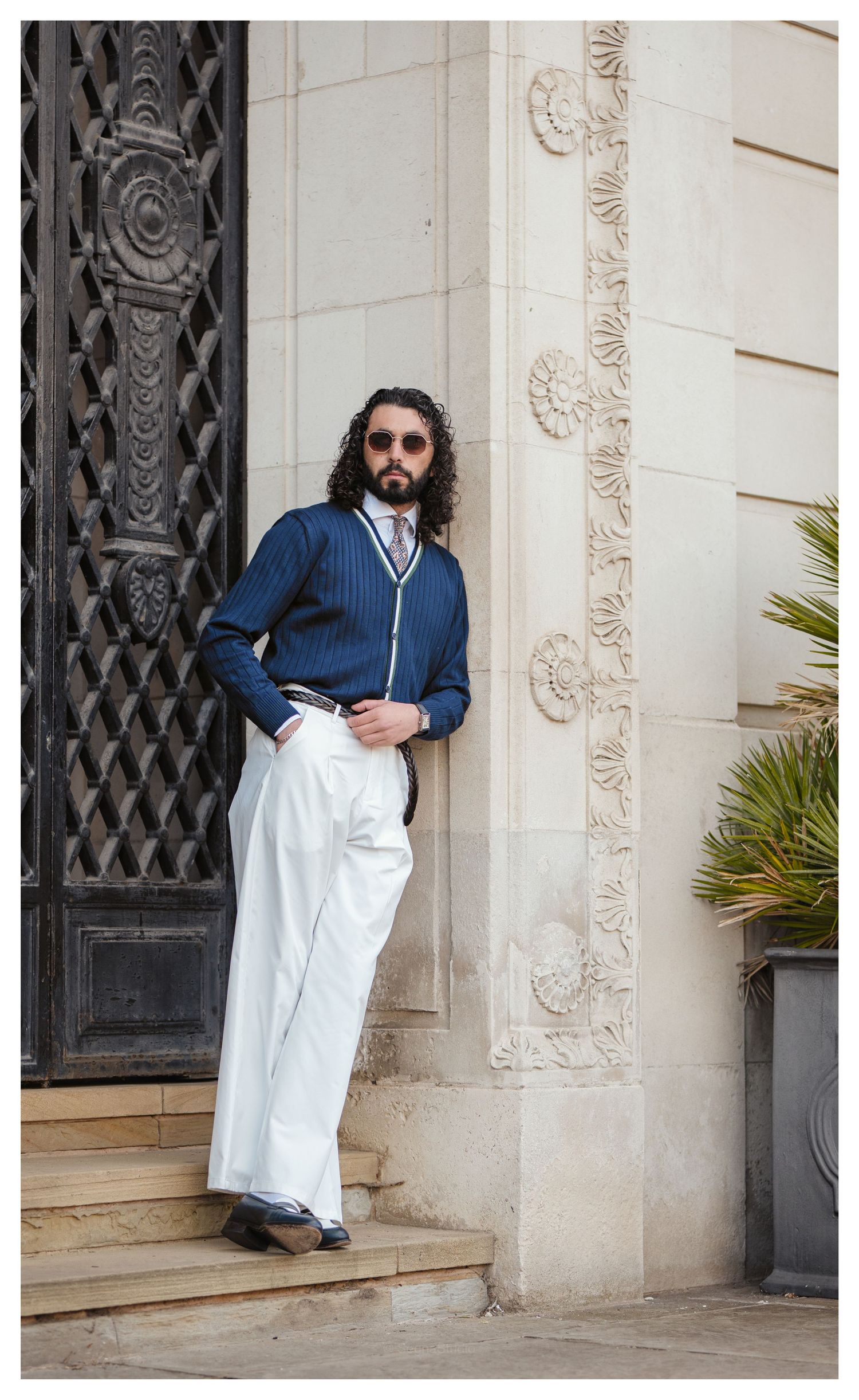 Stylish man in a navy cardigan and white trousers leaning against an ornate stone building.